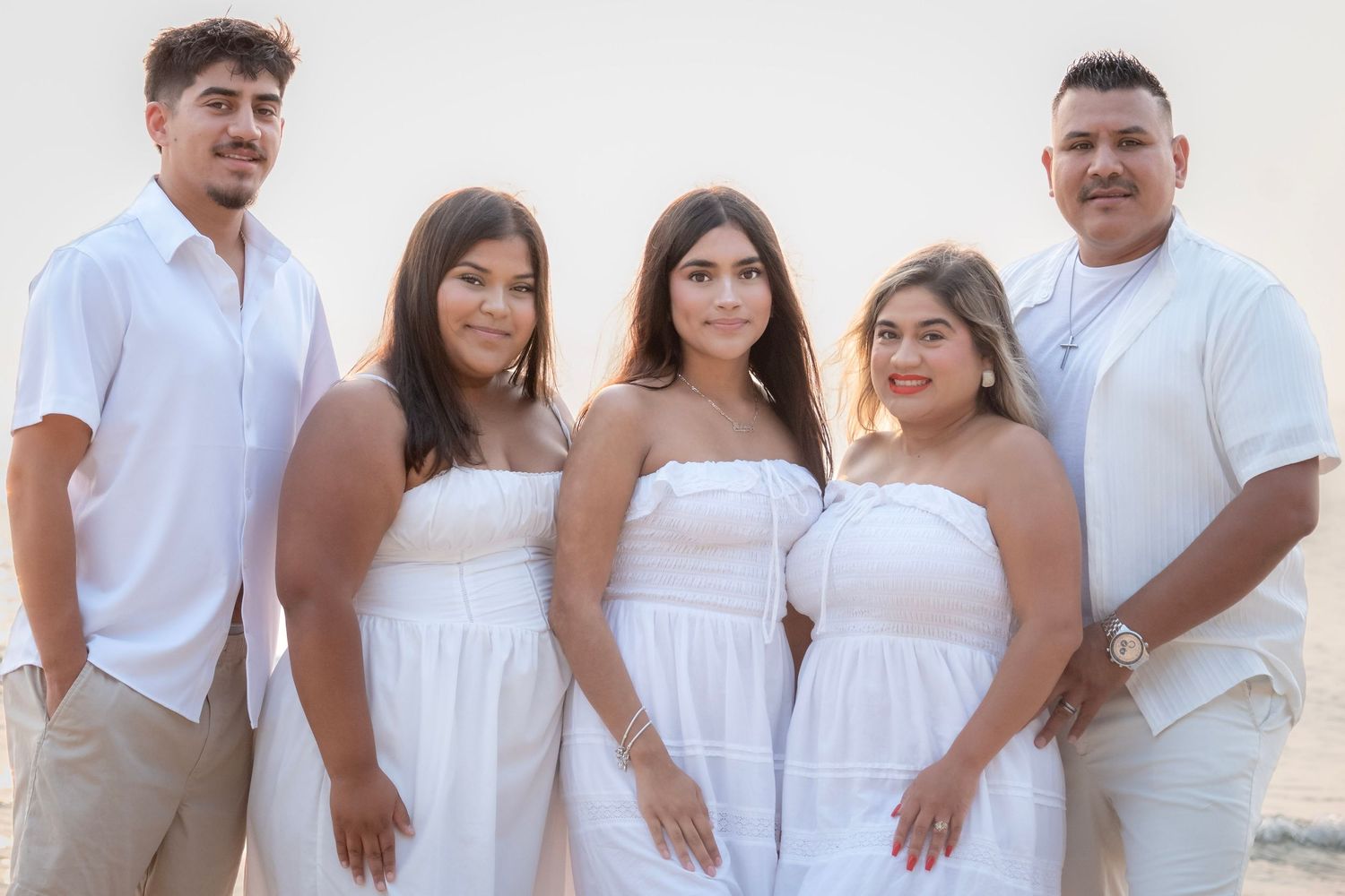 Five people in white outfits pose together on a beach at sunset for a family portrait photo.