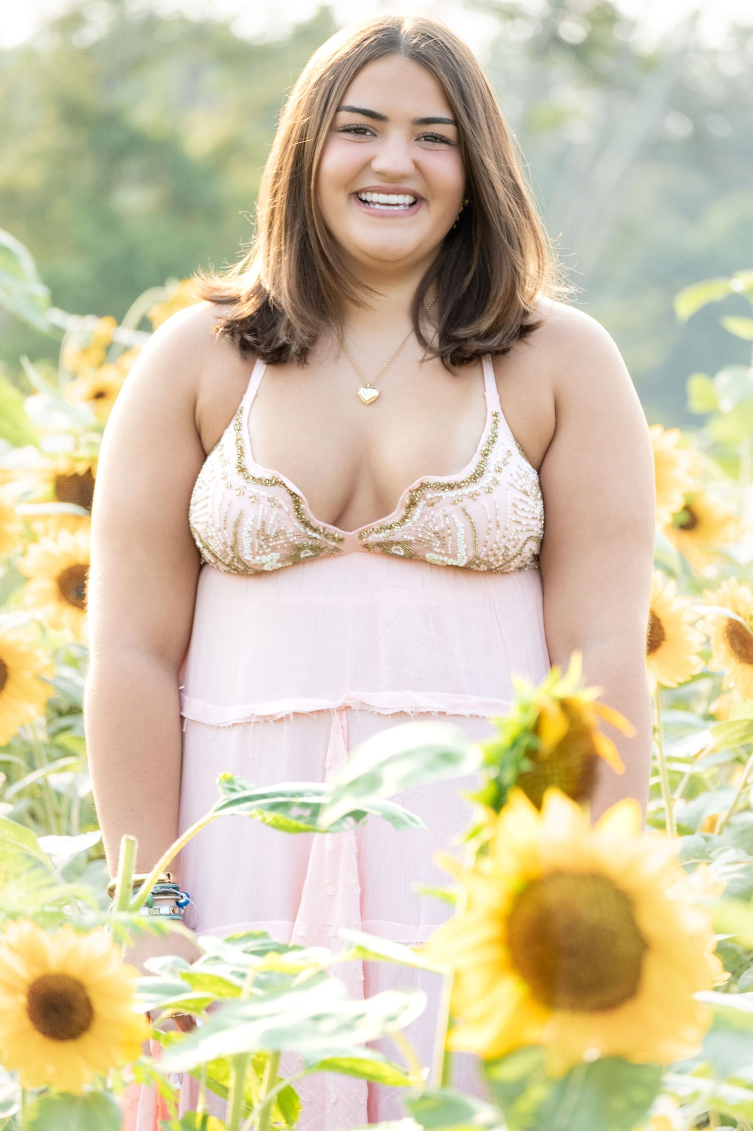 Smiling young woman in pink dress standing in a sunny sunflower field.