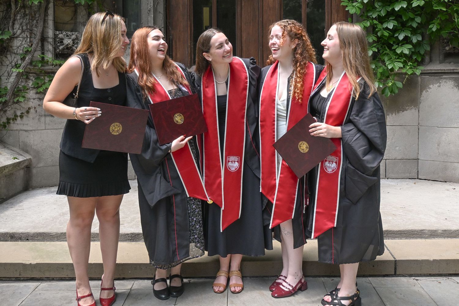 Five college graduates in caps and gowns celebrate together outdoors, holding diplomas and wearing red graduation stoles.