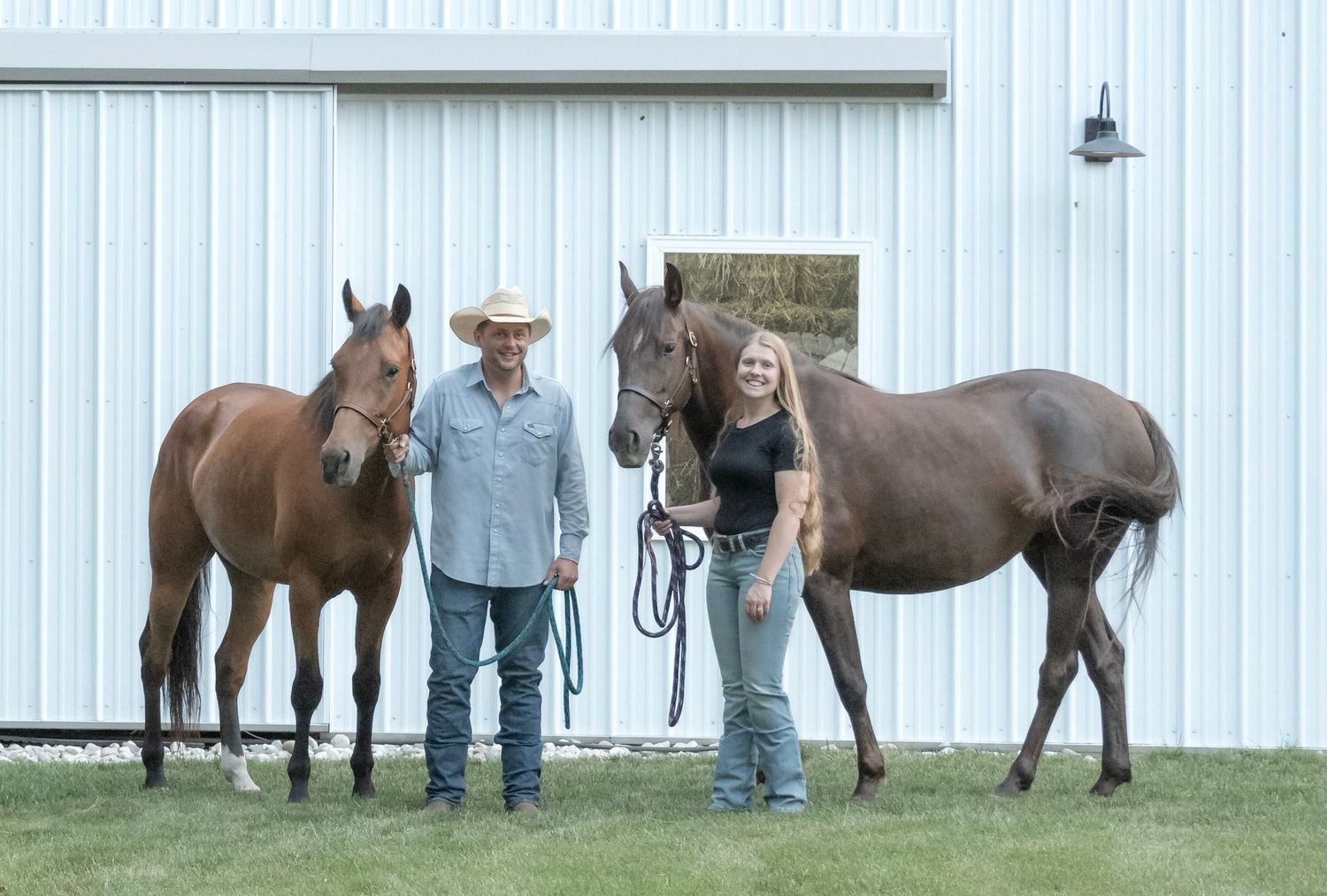 Man and woman standing between two brown horses outside a white barn on a sunny day.