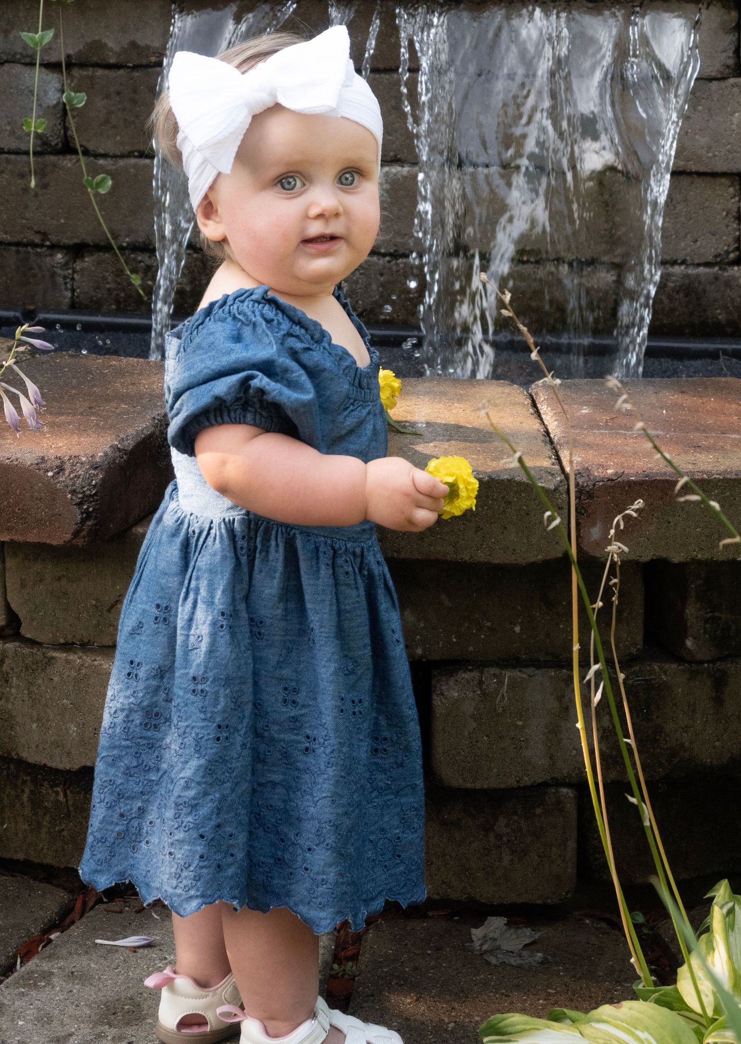 Toddler girl in blue dress and white bow headband holds yellow flowers near a stone fountain outdoors.