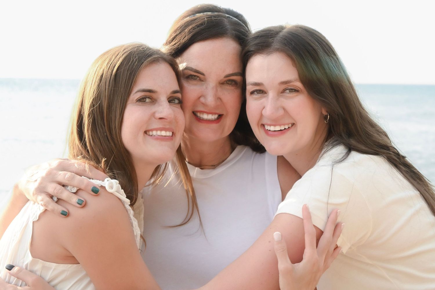 Three smiling women in white tops embrace on a sunny beach, sharing a joyful moment together.