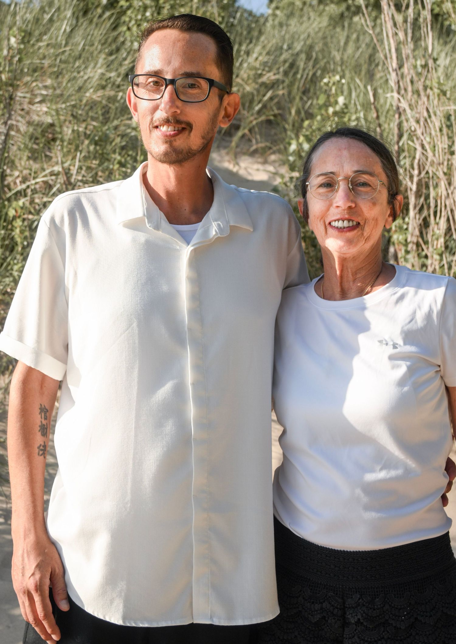 A young man wearing glasses and a white shirt stands beside an older woman in a white top outdoors.