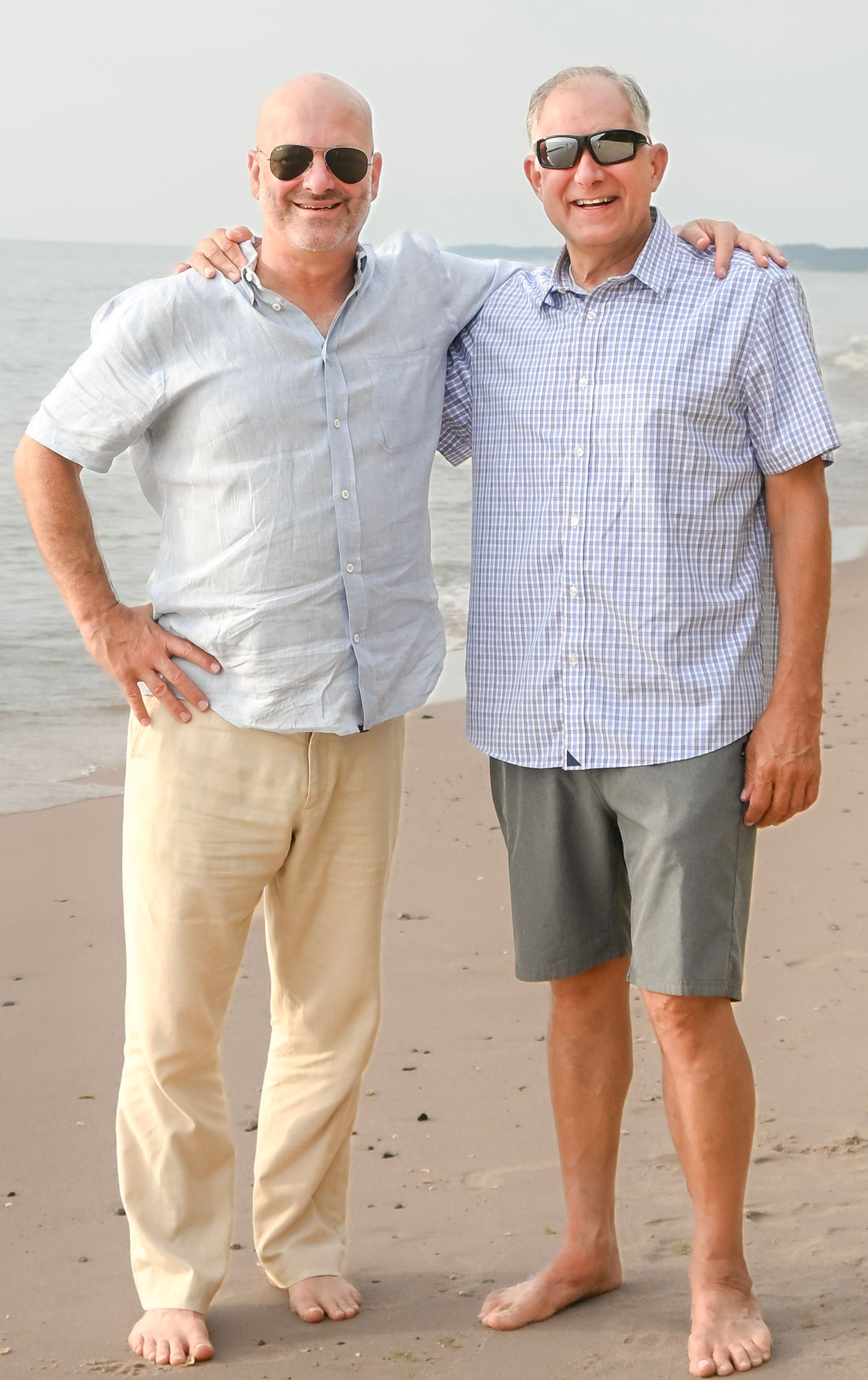 Two smiling men in sunglasses pose together barefoot on a sandy beach near the ocean.