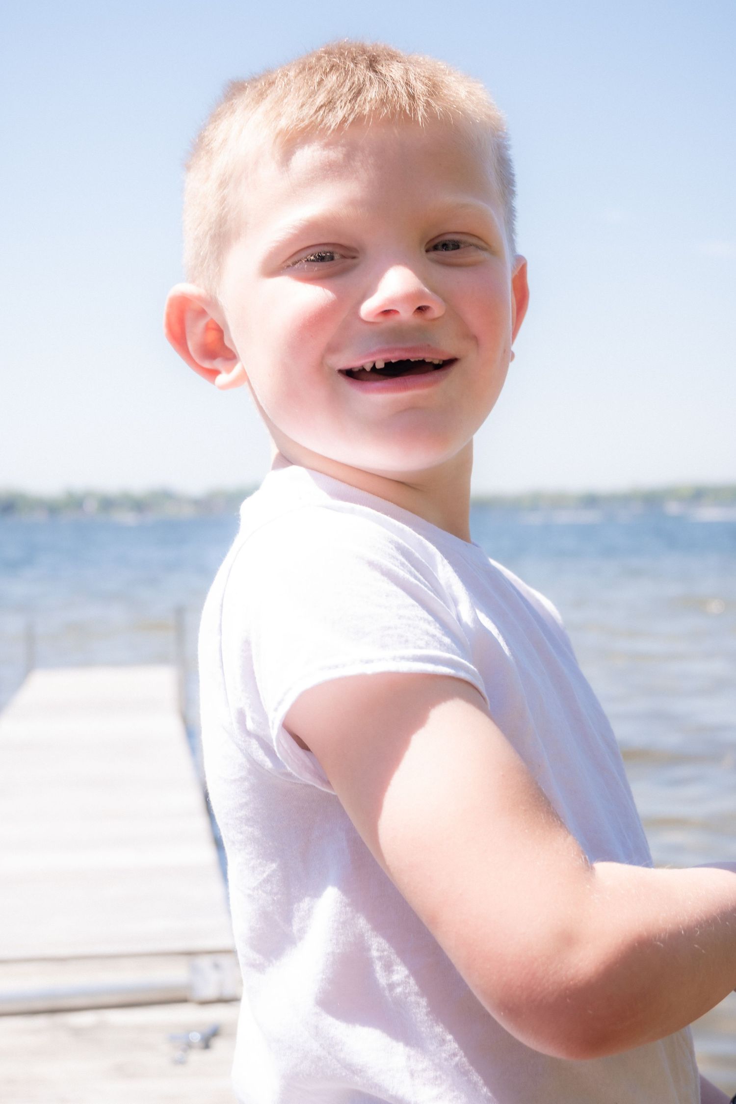 A cheerful young blond boy in a white shirt smiling on a sunny dock near the water.
