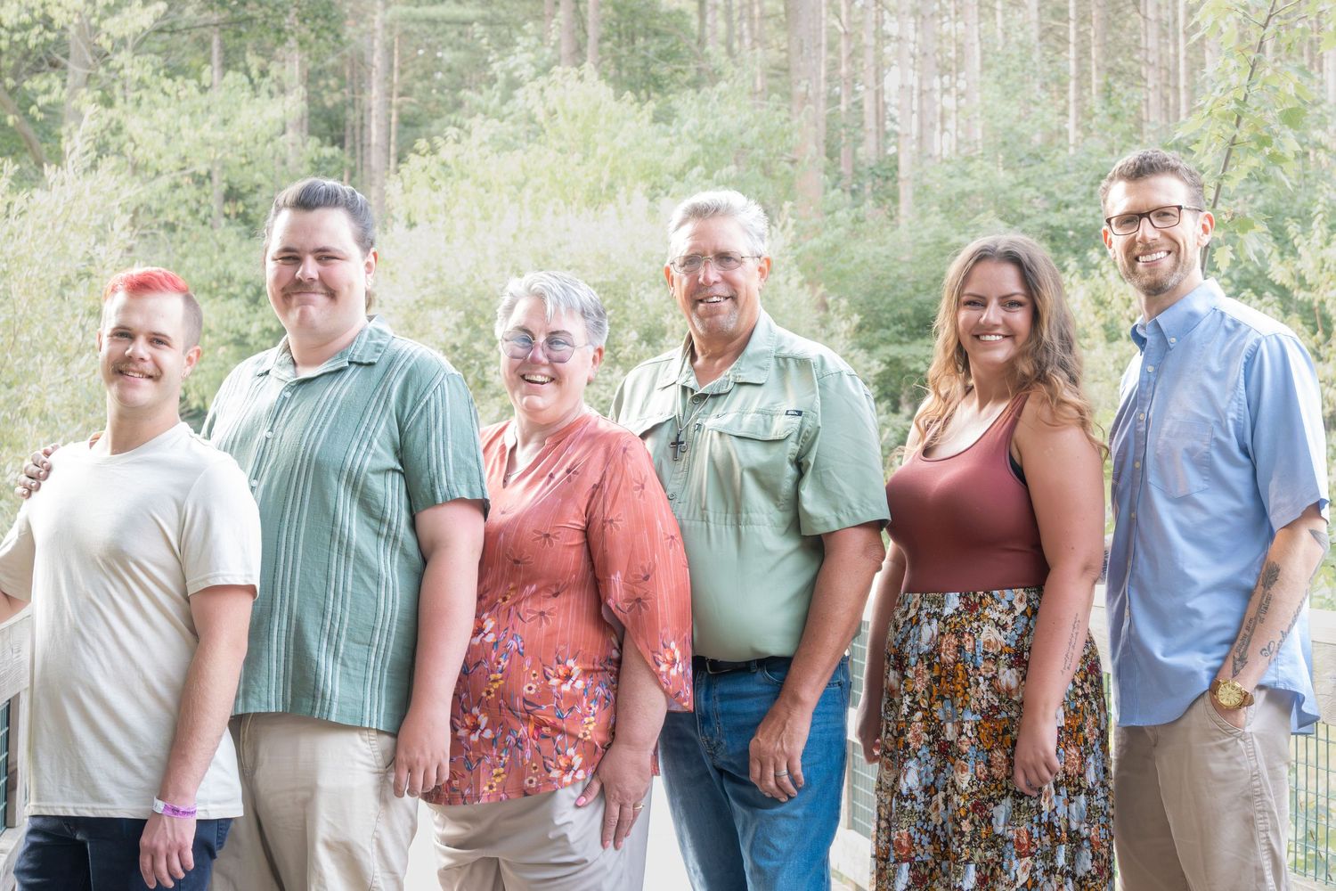 Six smiling adults pose together outdoors in a wooded setting, wearing casual summer attire for a family group photo.