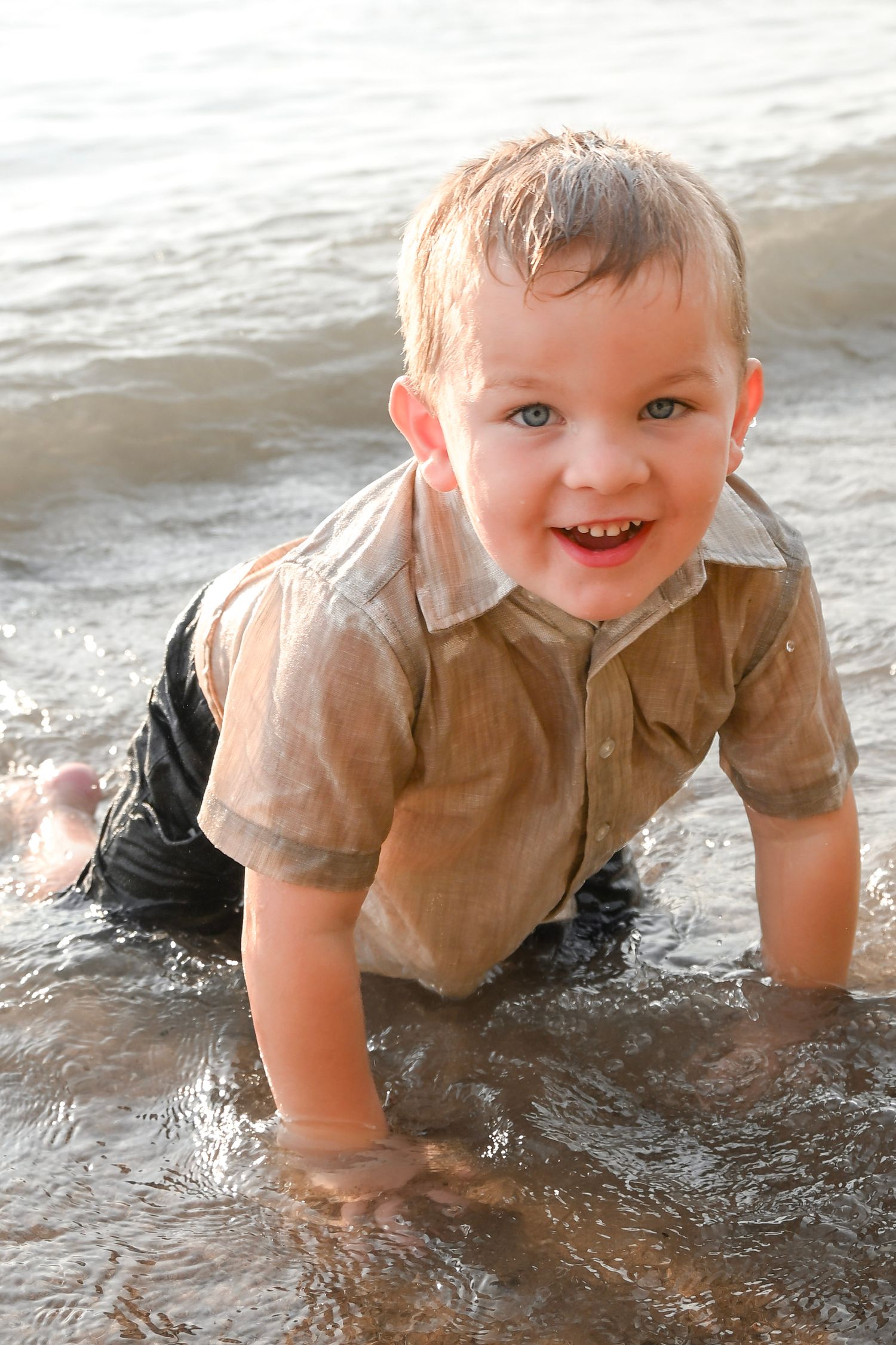 Happy toddler boy playing in ocean waves on the beach, soaking wet in a button-up shirt, smiling joyfully.