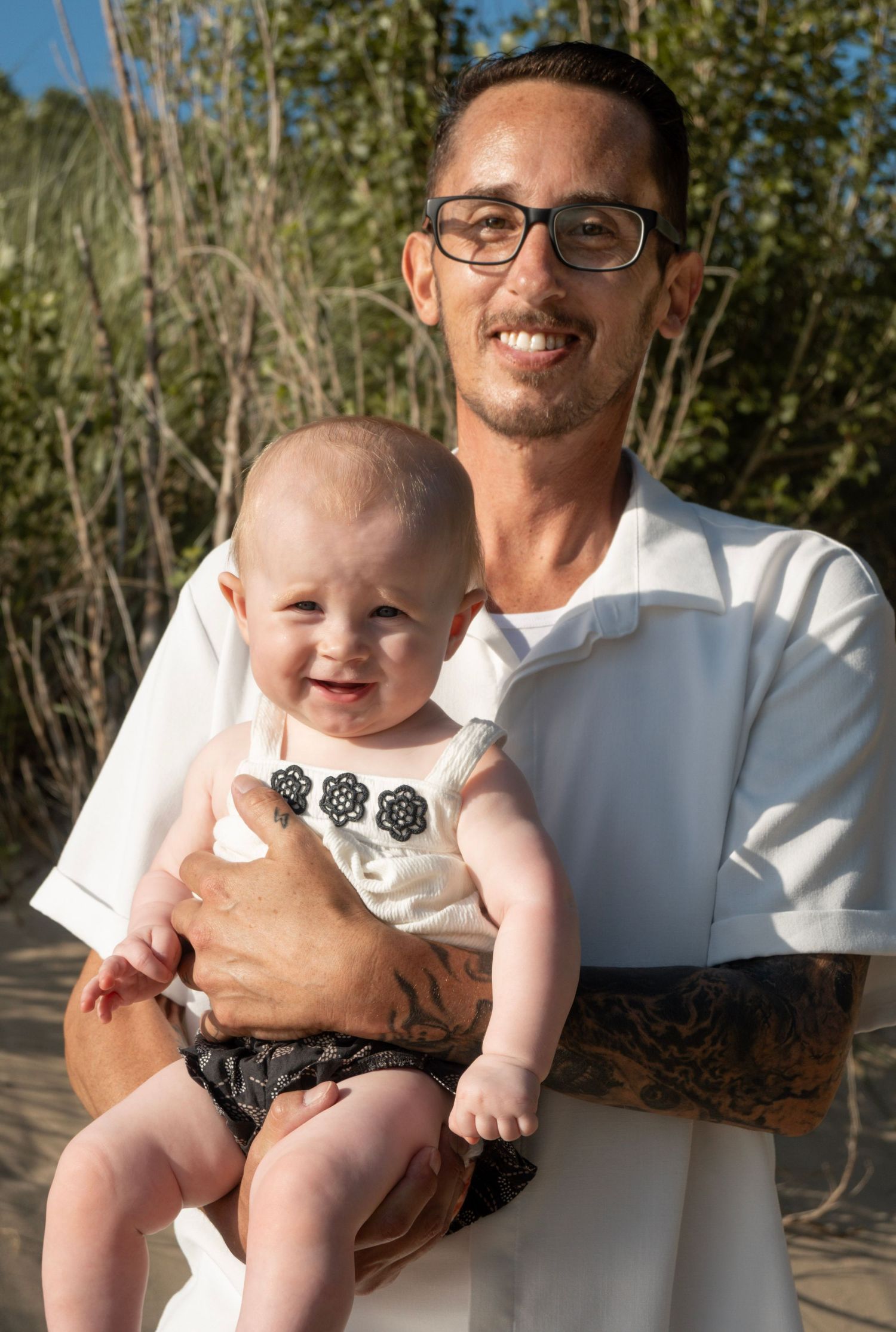 Smiling man with glasses and tattoos holding a happy baby outdoors near sandy dunes and tall green plants.