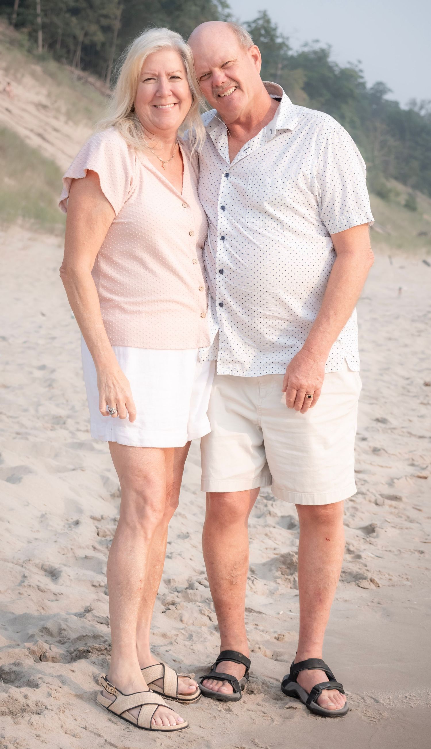 Smiling senior couple in white summer outfits posing together on a sandy beach with trees in the background.