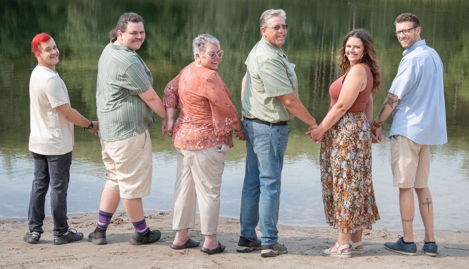 Six people pose together outdoors near a lake shore, smiling for a family portrait on a sunny day.