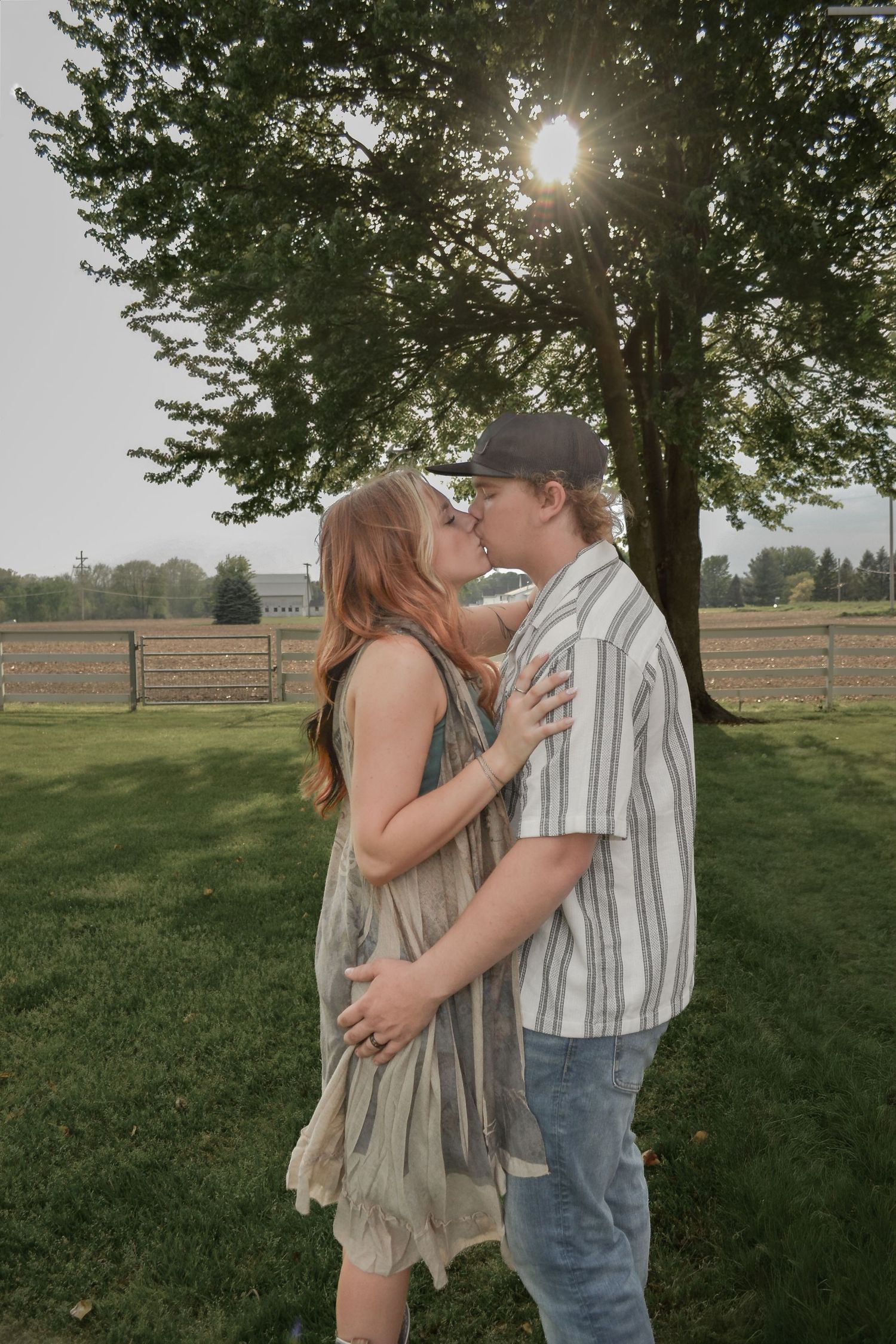 Couple sharing a romantic kiss outdoors under a large tree with sunlight streaming through the branches.