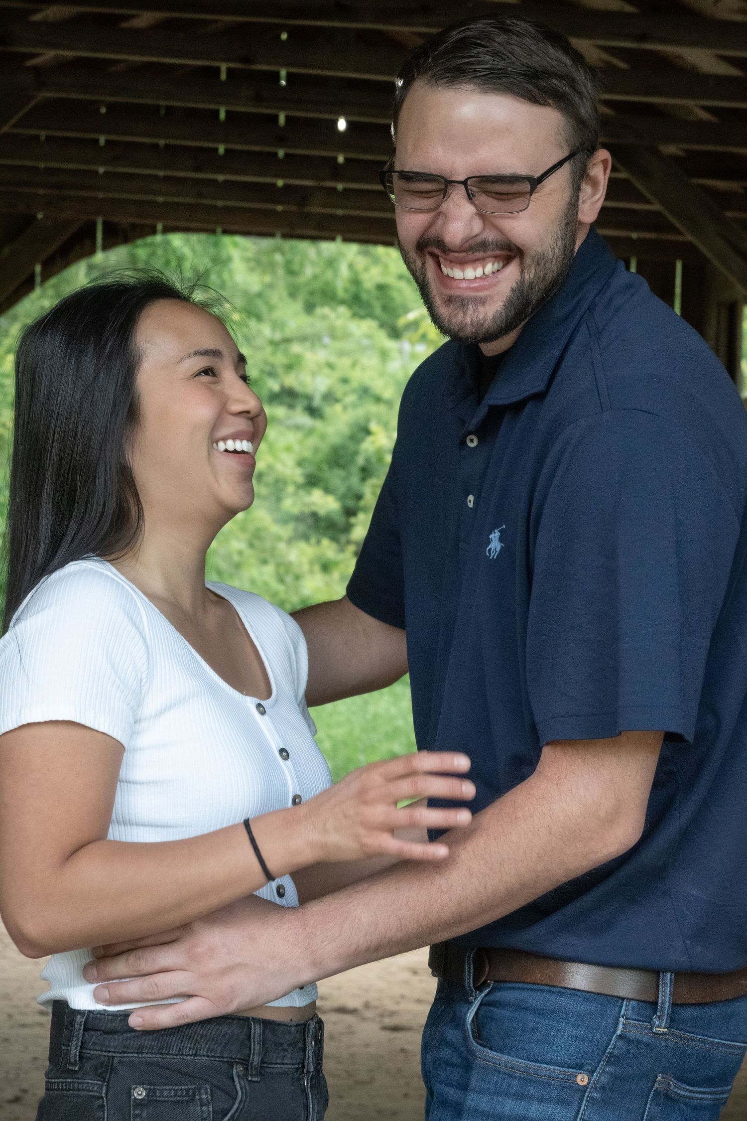 Laughing couple embracing and sharing a joyful moment under a covered wooden bridge in a park.