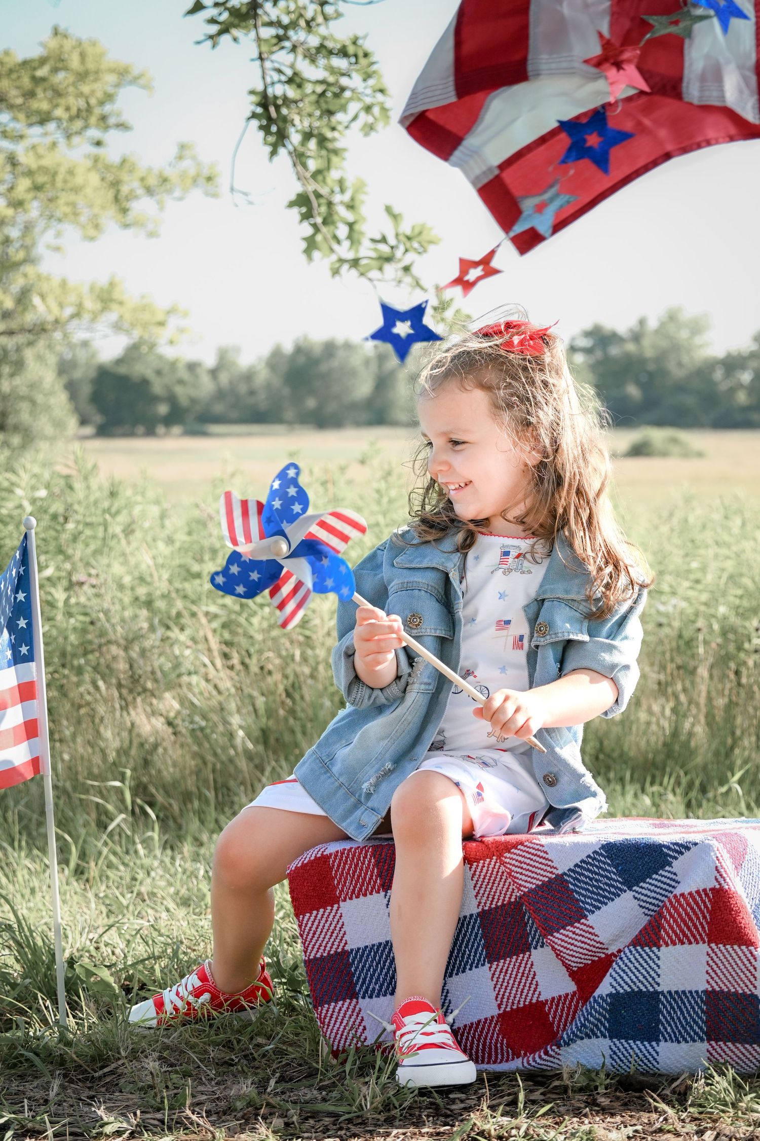 Young girl sitting outdoors holding a patriotic pinwheel, surrounded by American flags on Independence Day.