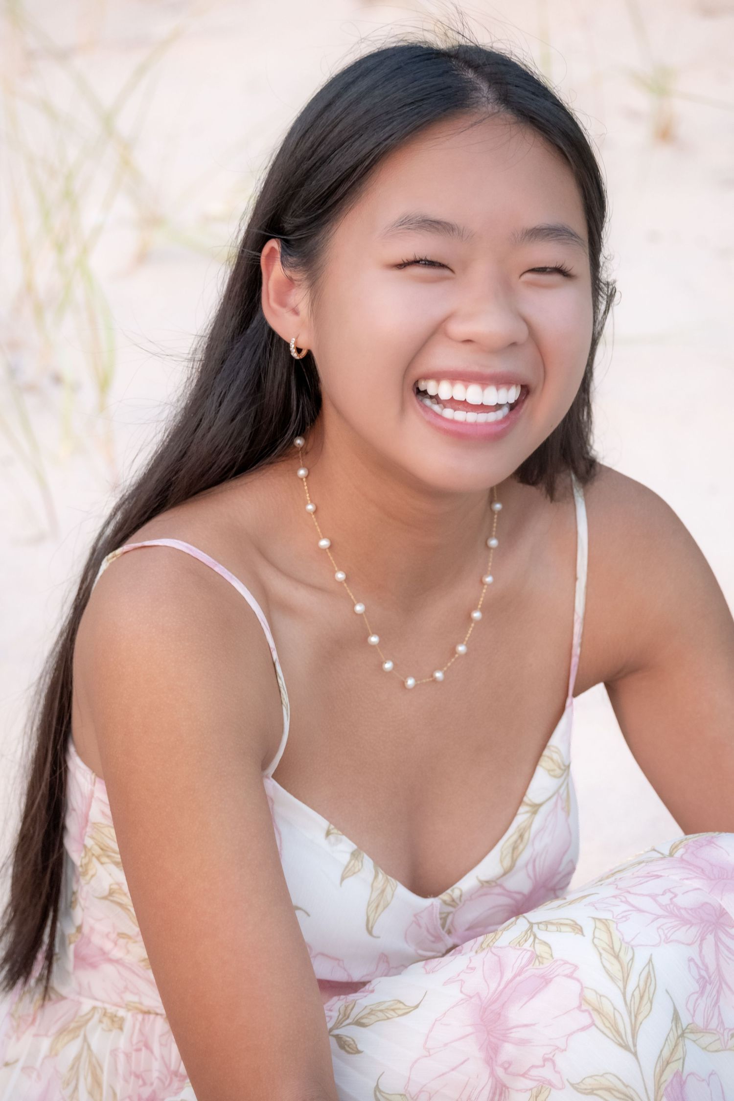 Young Asian woman laughing joyfully on a sandy beach, wearing a floral dress and pearl necklace.