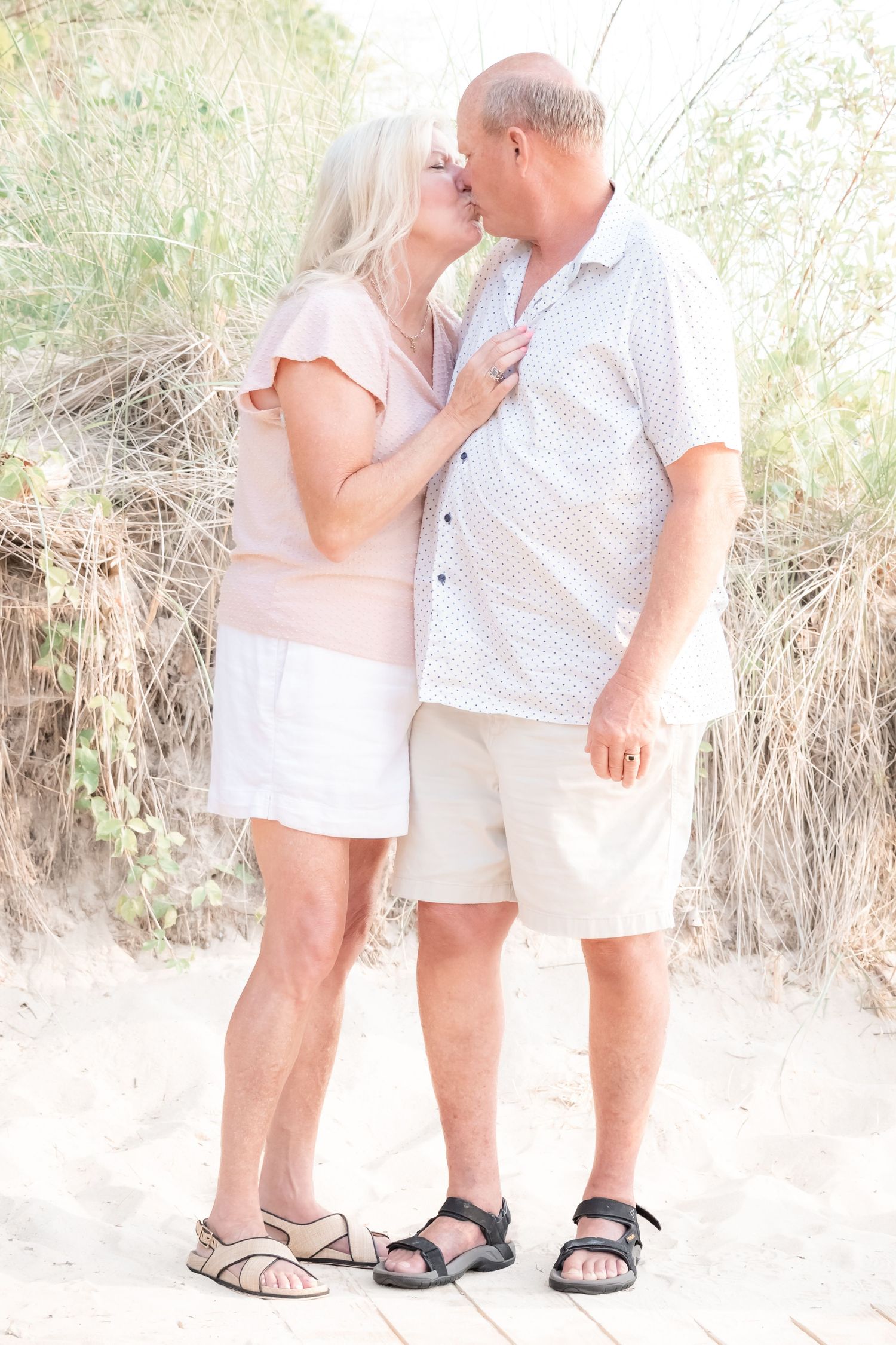 Older couple sharing a romantic kiss on sandy beach, wearing casual summer clothes and sandals.
