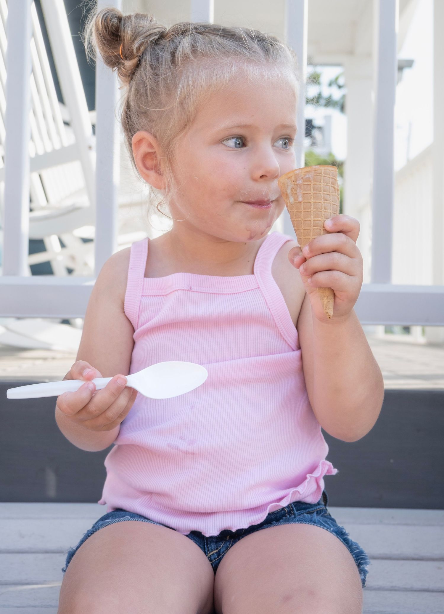 Young toddler girl in pink tank top enjoying an ice cream cone while holding a spoon outdoors on a sunny day.