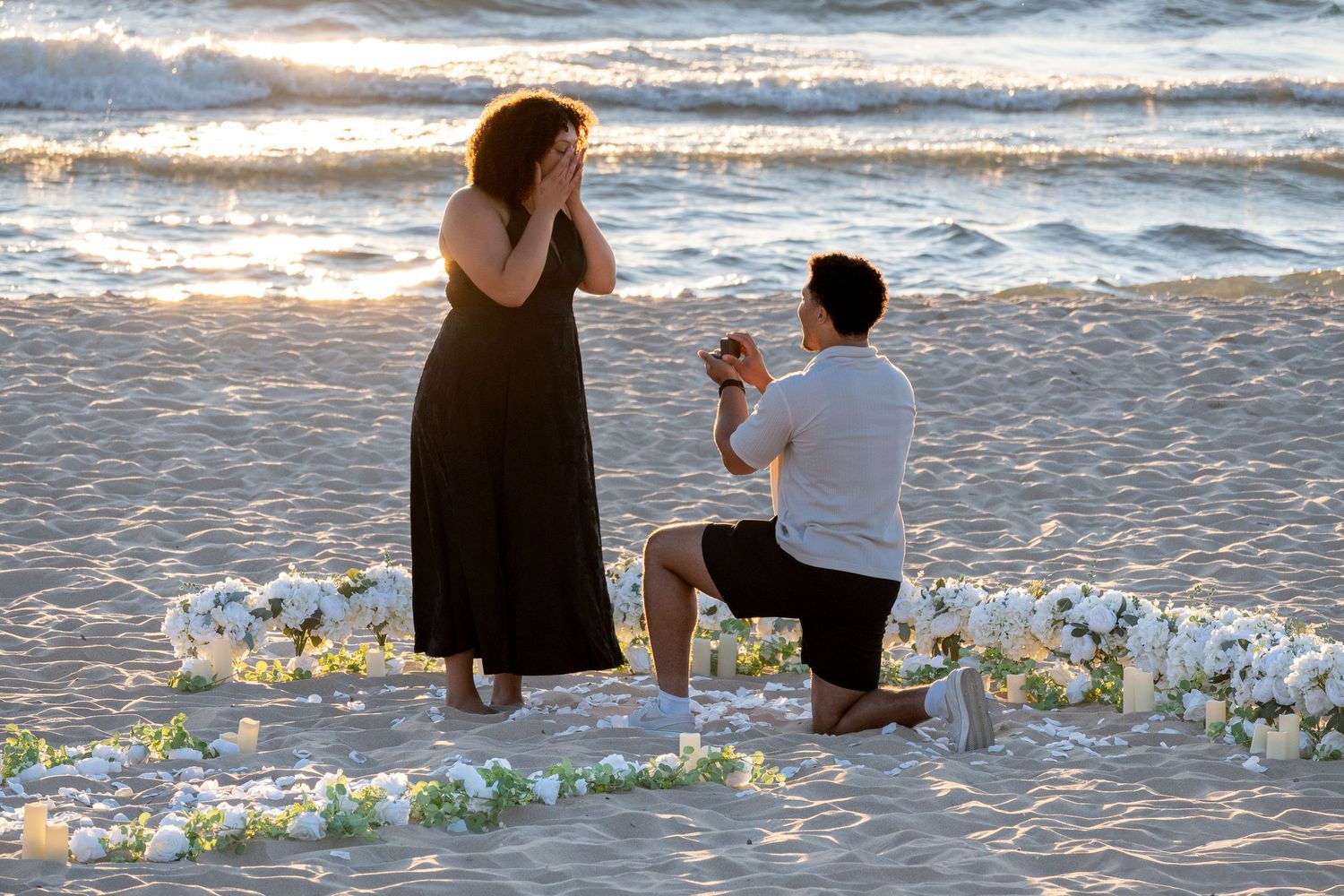 Man proposes to woman on beach at sunset surrounded by white flowers arranged in a heart shape.