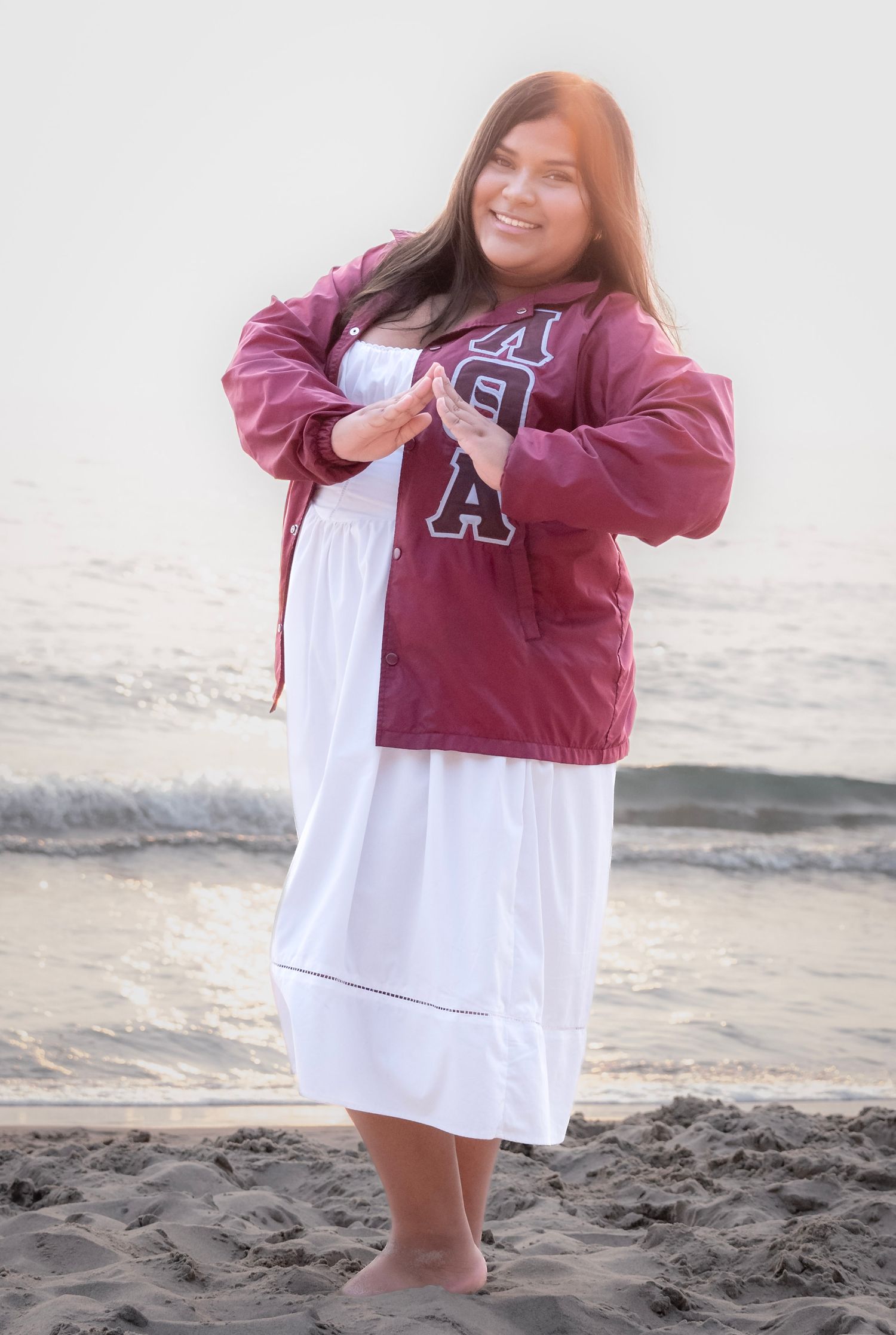 Smiling woman in maroon jacket and white skirt posing barefoot on a sandy beach near ocean waves.