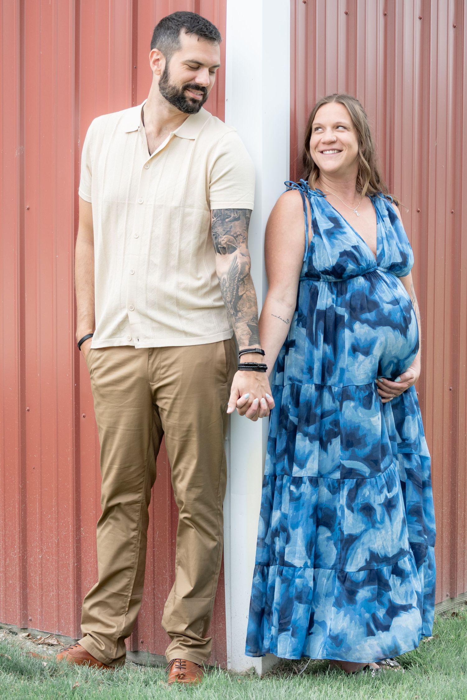 Smiling pregnant couple holding hands against red barn wall, man in beige shirt and khakis, woman in blue maxi dress.