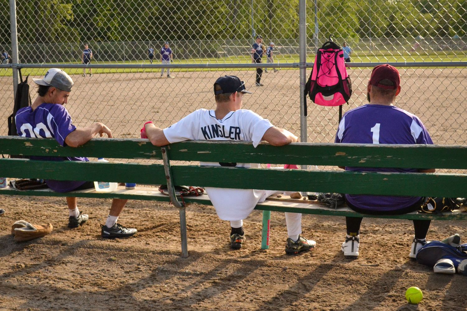 Three softball players sitting on a green dugout bench watching a game on a sunny day.
