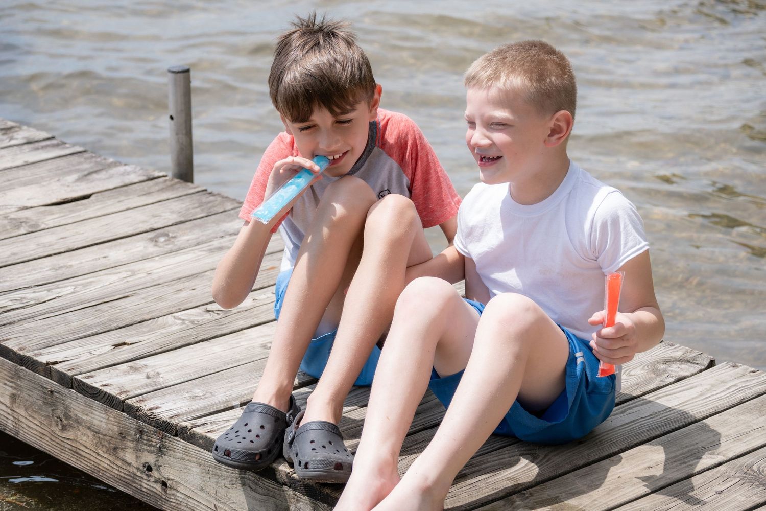 Two boys sitting on a wooden dock eating colorful popsicles by the water on a sunny day.