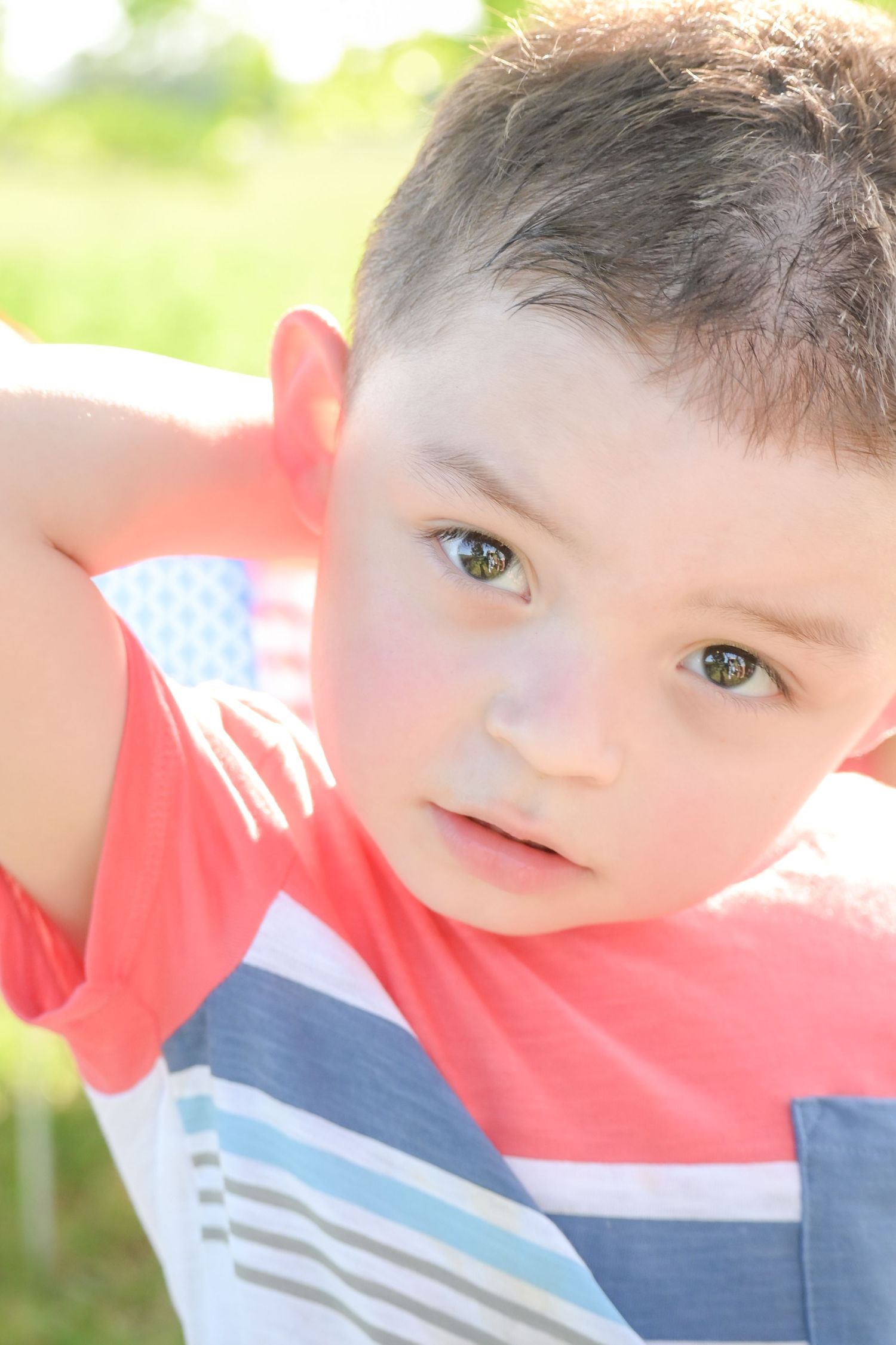 Young boy in striped shirt with American flag in background outdoors.
