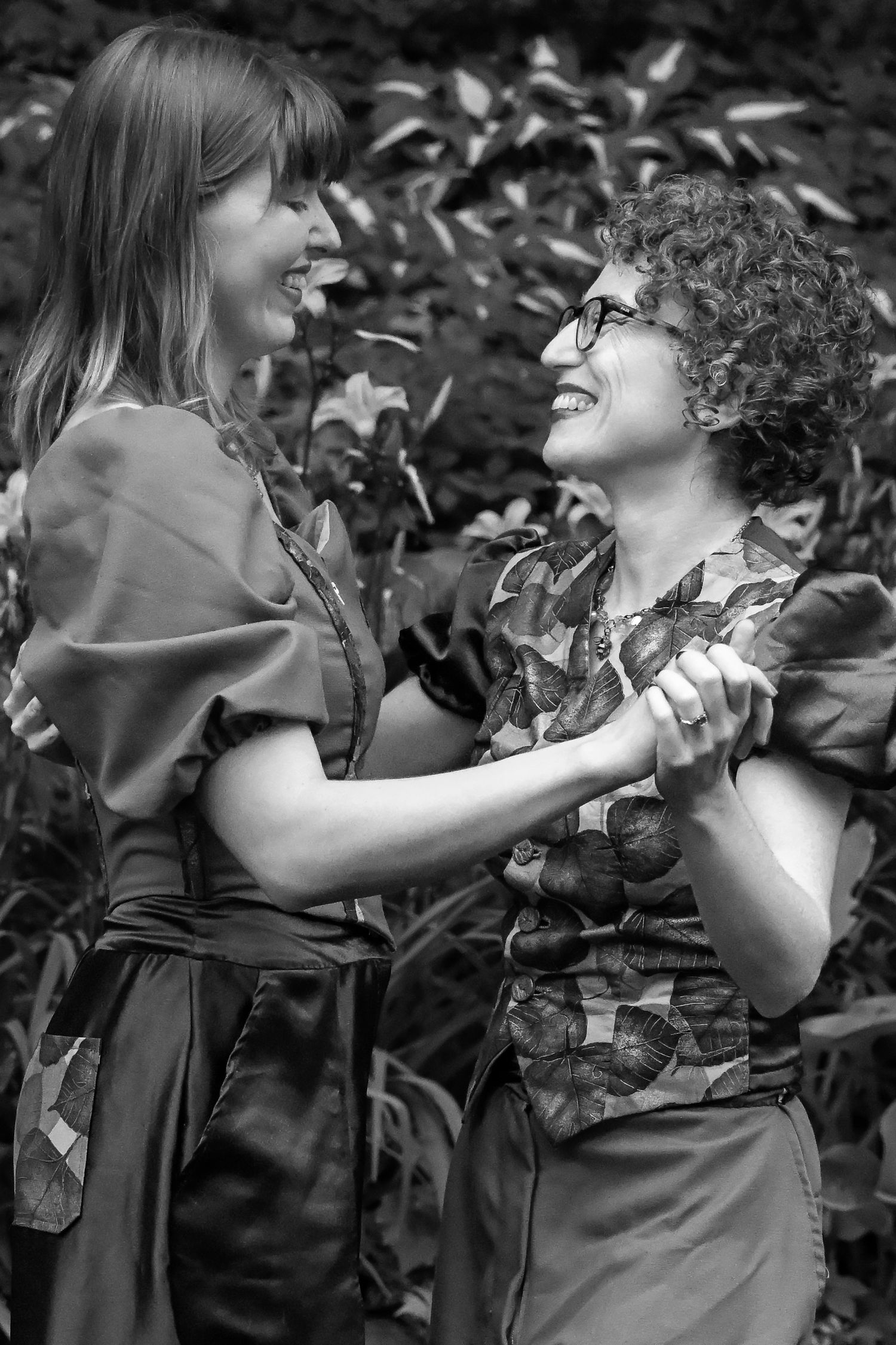 Two women smiling and holding hands outdoors among lush foliage, captured in black and white photography.