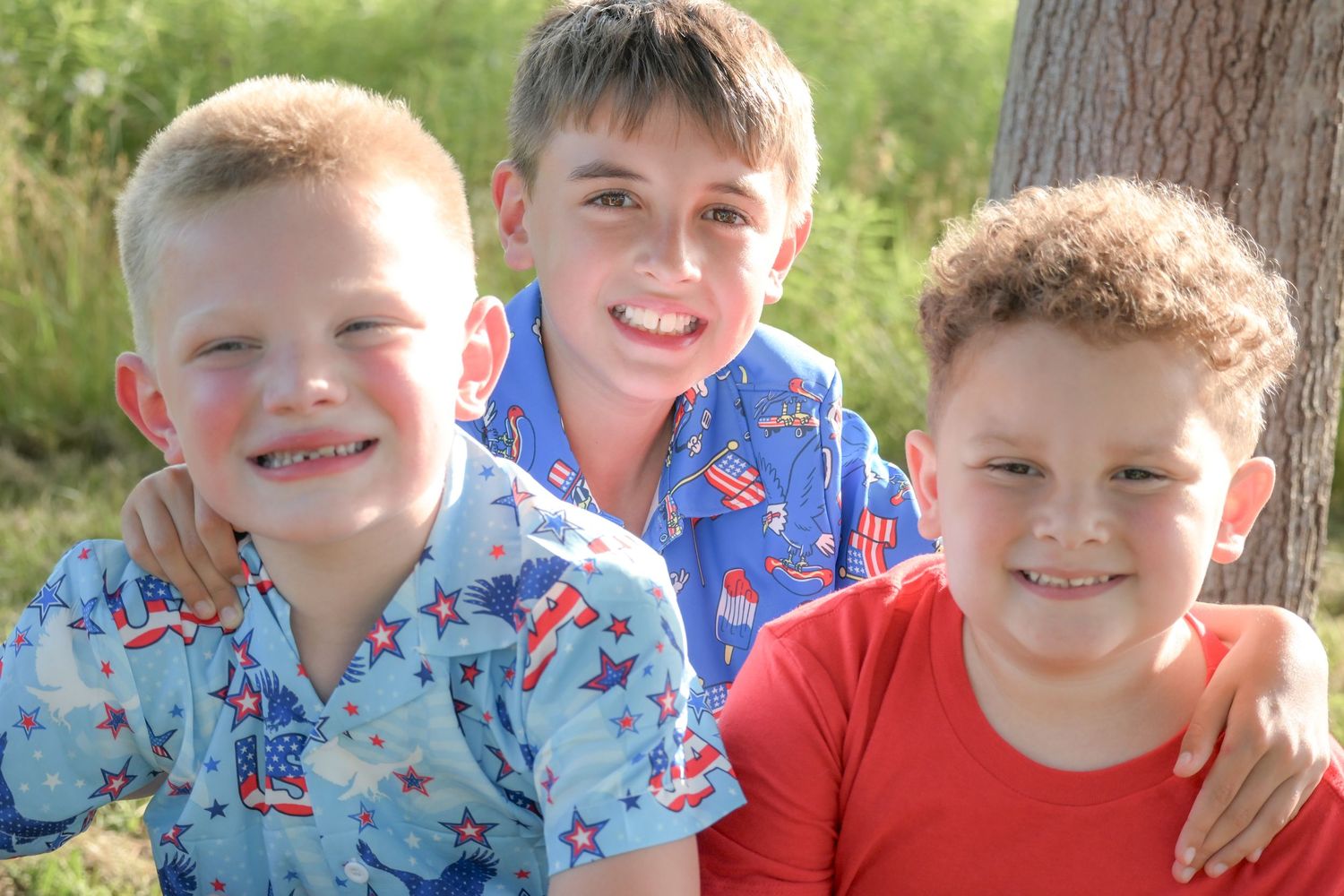 Three smiling boys in patriotic shirts pose together outdoors near a tree on a sunny day.