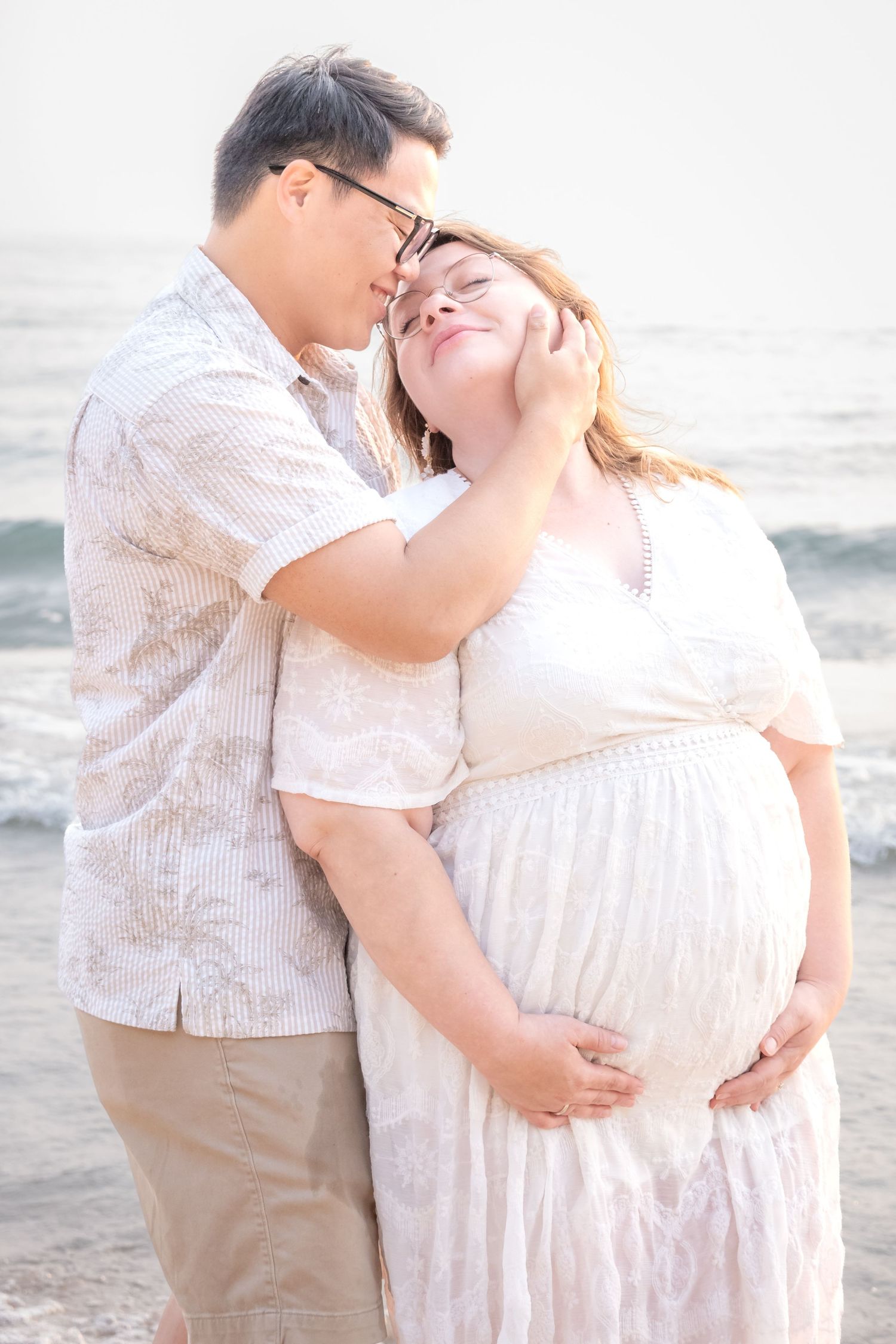 Couple sharing tender moment on beach, pregnant woman in white dress cradled by partner near ocean waves.