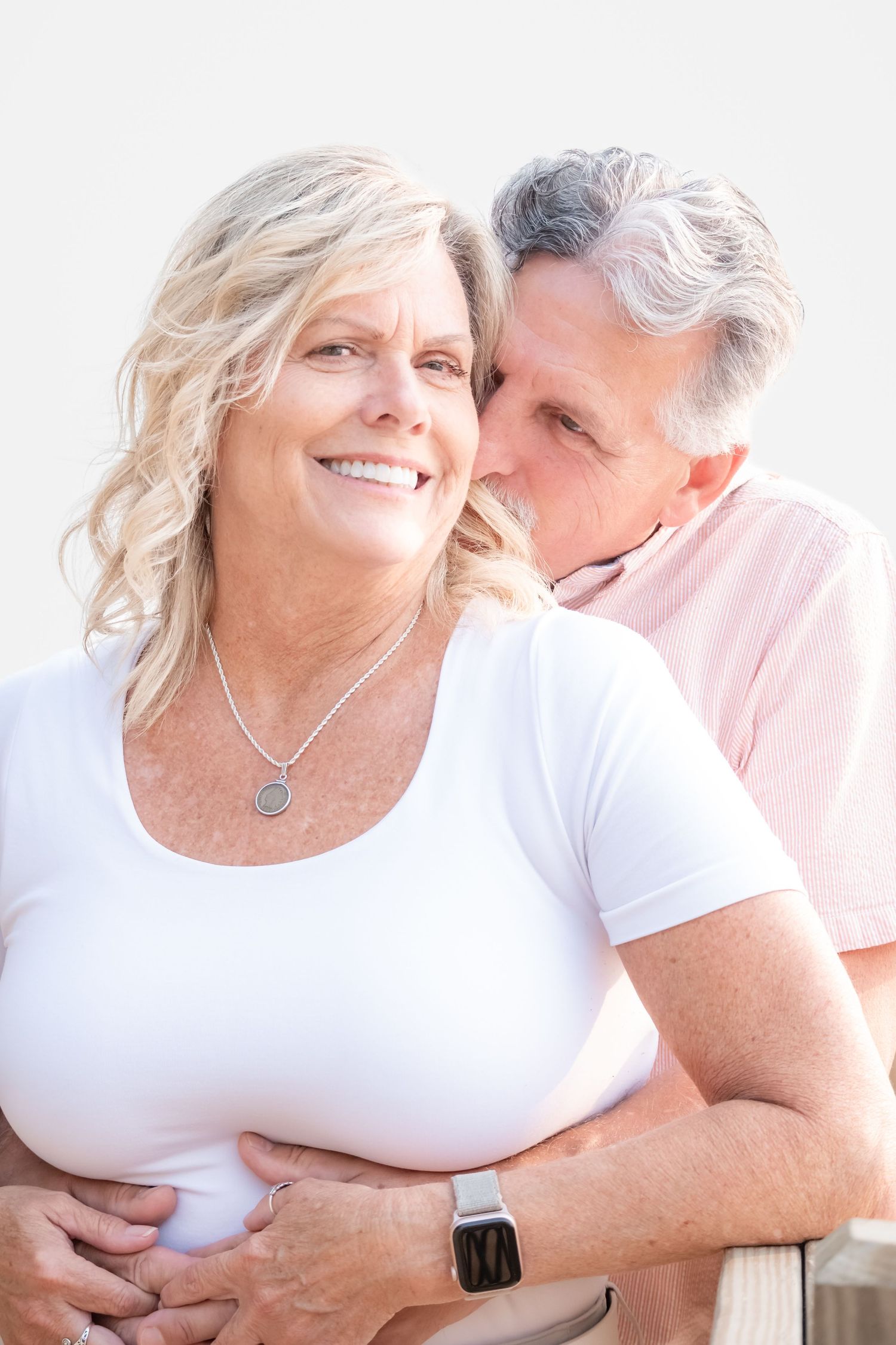 Smiling middle-aged blonde woman in white top embraced from behind by gray-haired man whispering in her ear.