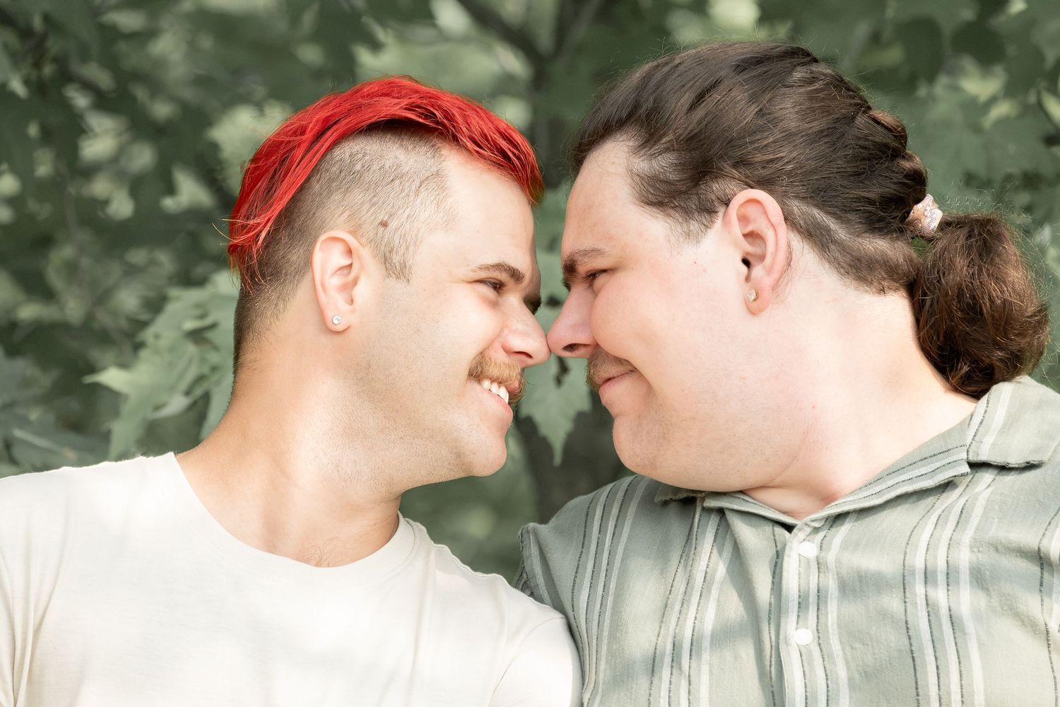 Two men with colorful hair share a close romantic moment outdoors surrounded by green foliage.