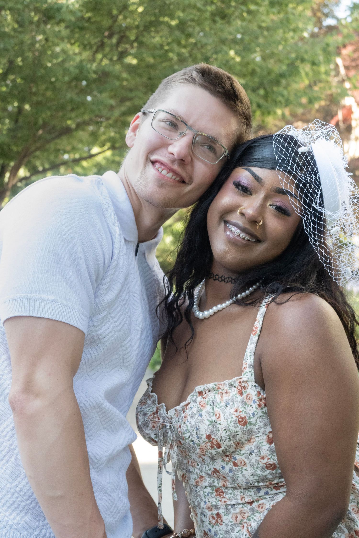 Happy interracial couple posing outdoors, bride wearing floral dress and veil, groom in white shirt, smiling together.