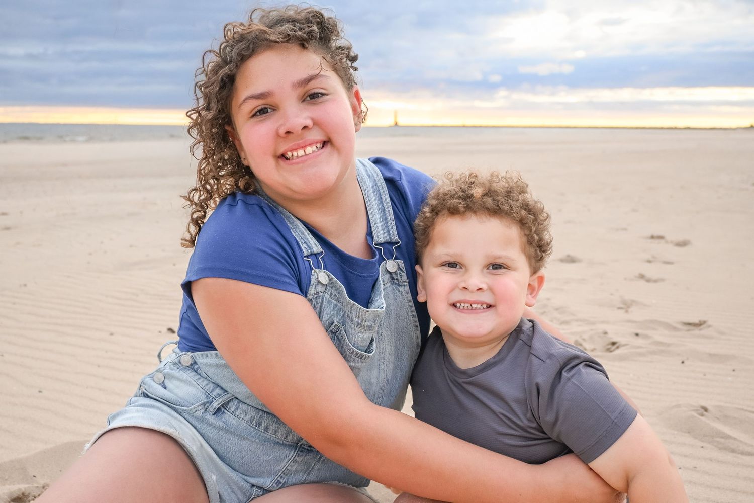 Two smiling children sitting together on a sandy beach at sunset, girl in denim overalls hugging younger boy.