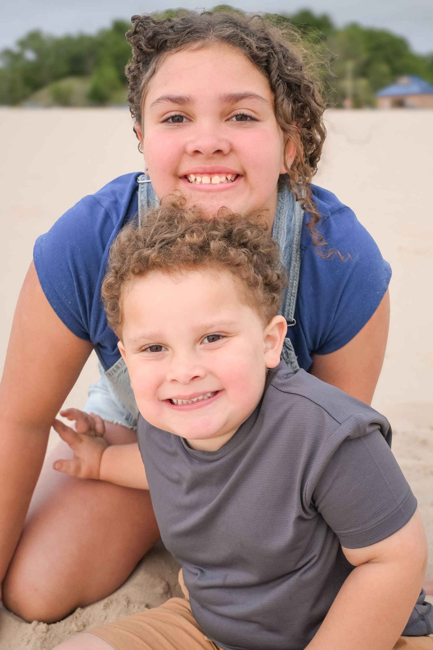 A smiling girl in denim overalls hugs a curly-haired boy on a sandy beach.