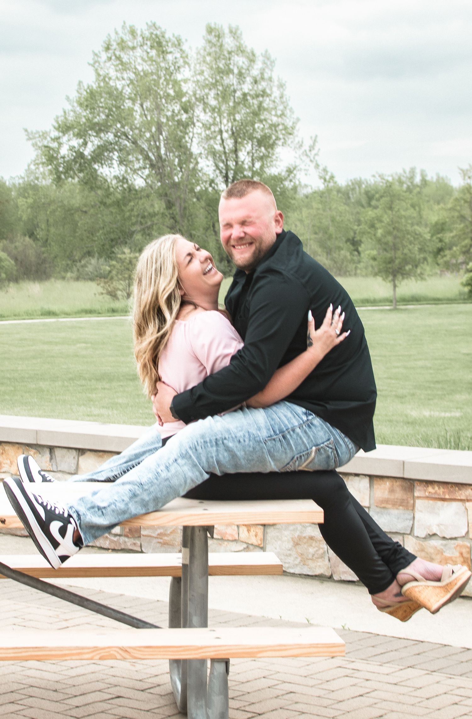Happy couple laughing and embracing while sitting on an outdoor bench surrounded by greenery.