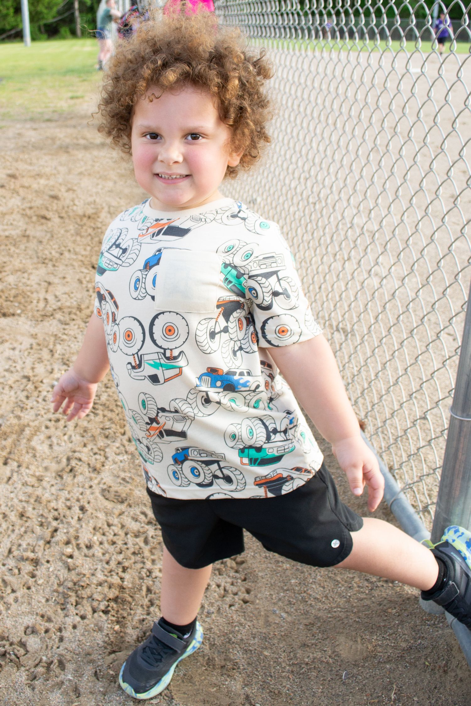 Curly-haired child smiling in a monster truck print shirt and black shorts near a chain-link fence outdoors.