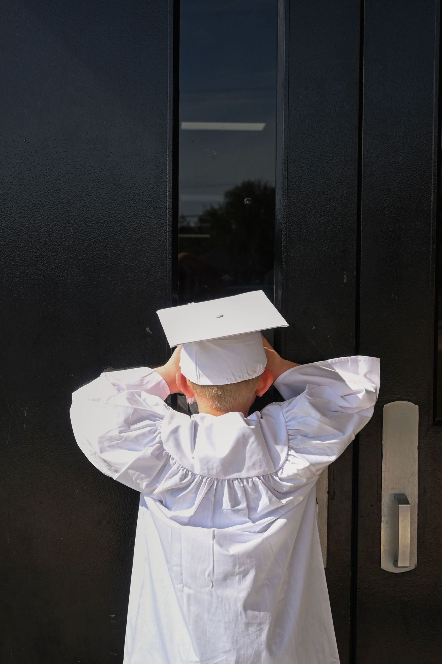 Graduate in white cap and gown stands facing black door with hands behind head.