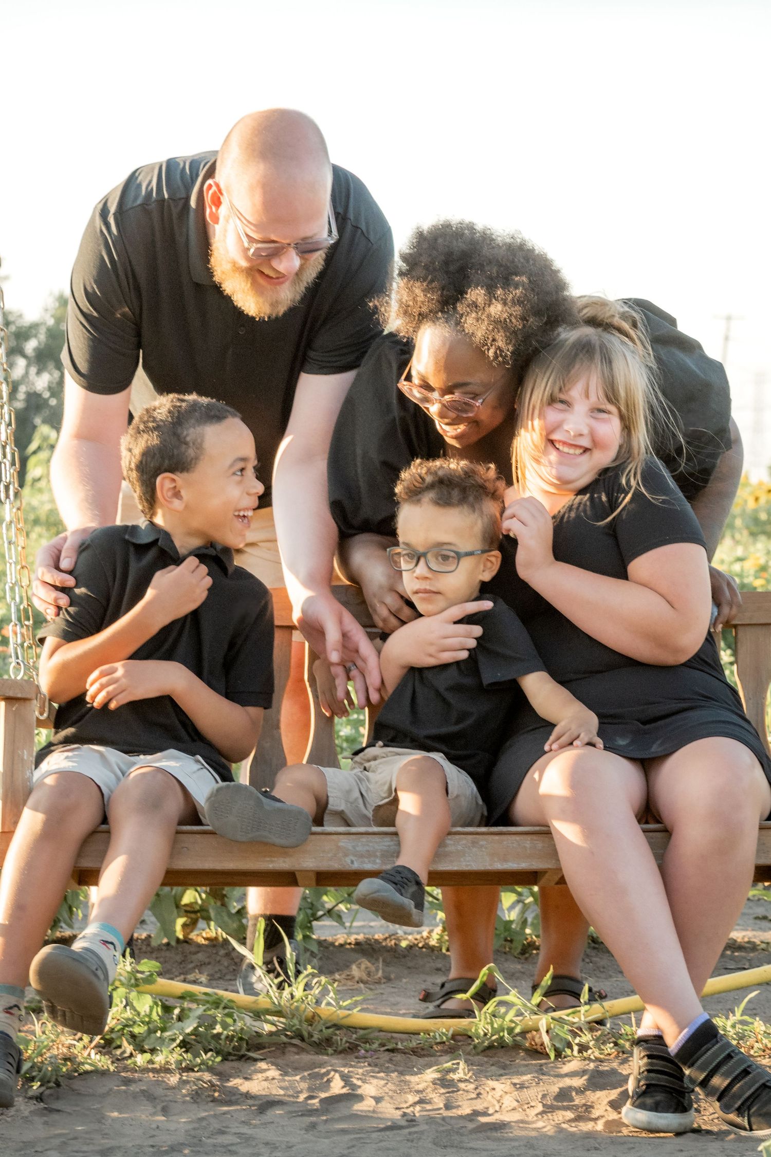 A joyful multiracial family of five in matching black outfits poses together on a wooden bench outdoors.
