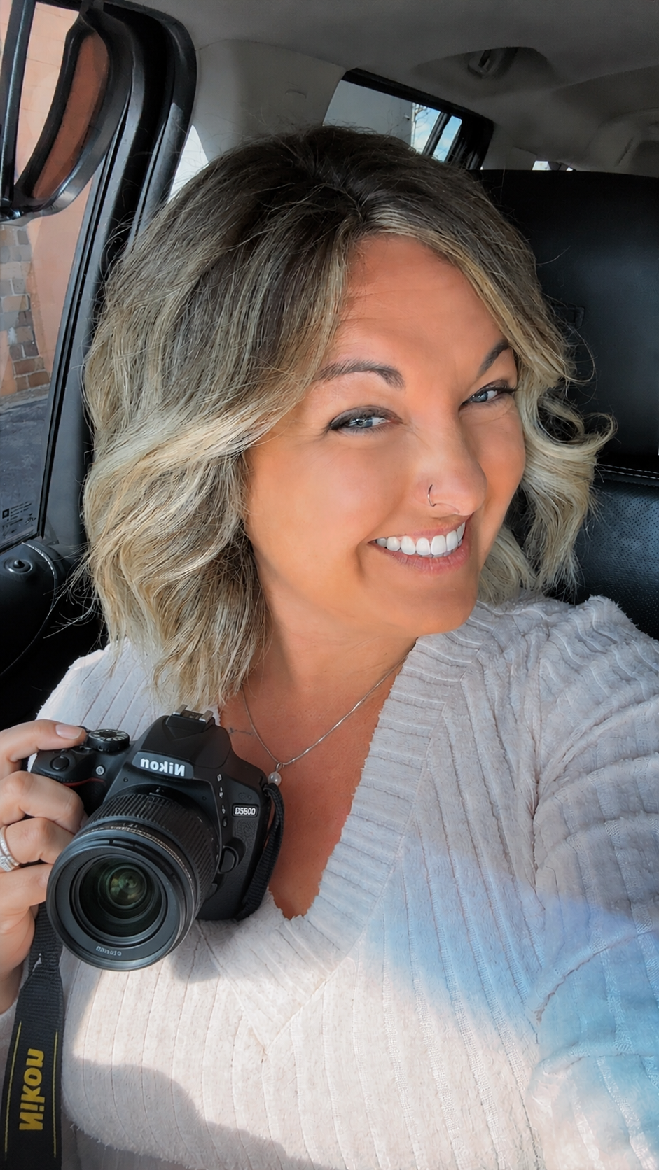 Smiling woman with blonde highlights holds a Nikon DSLR camera while sitting in a car.
