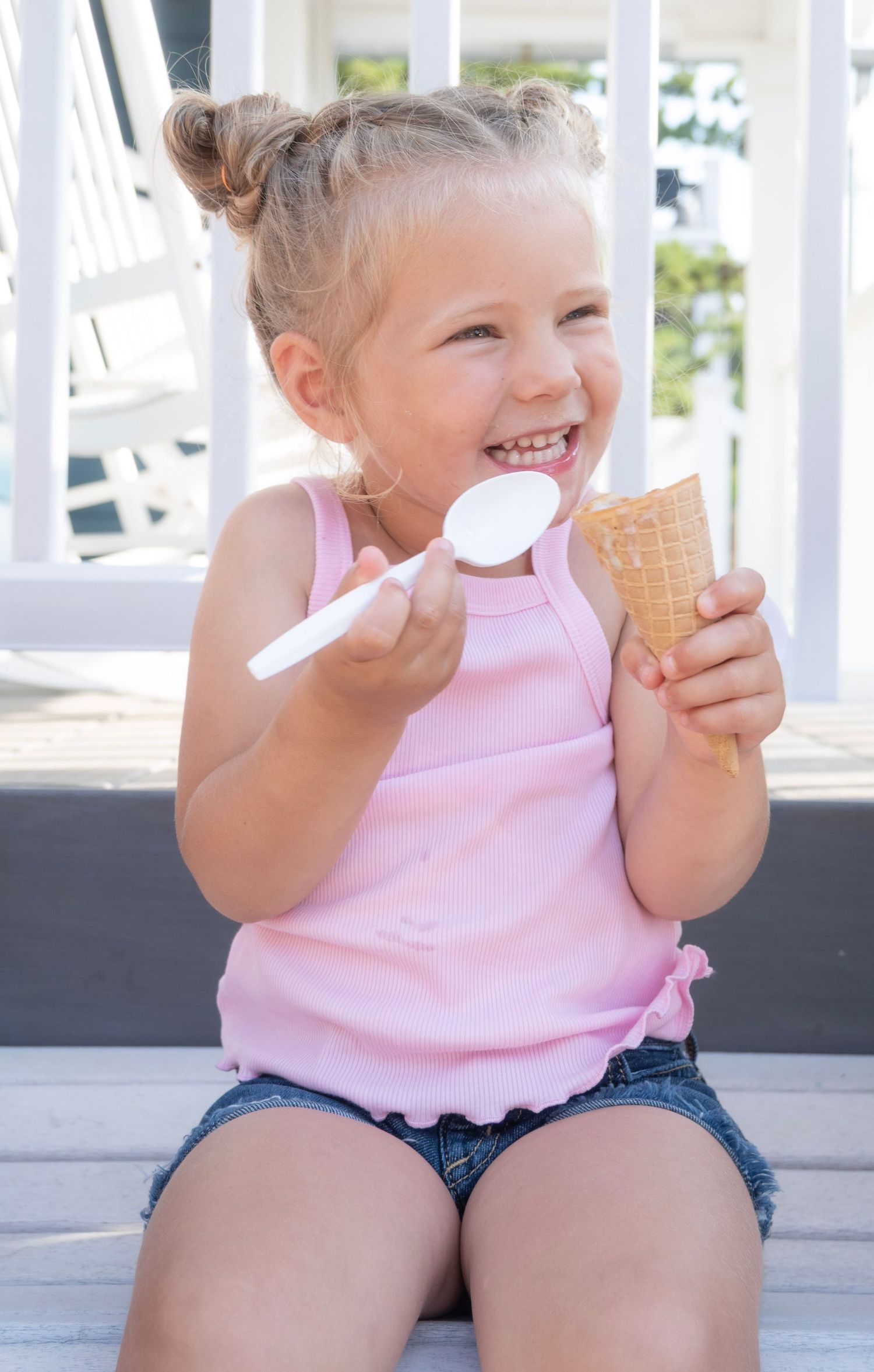 A happy young girl in a pink tank top enjoys an ice cream cone with a spoon outdoors on a sunny day.