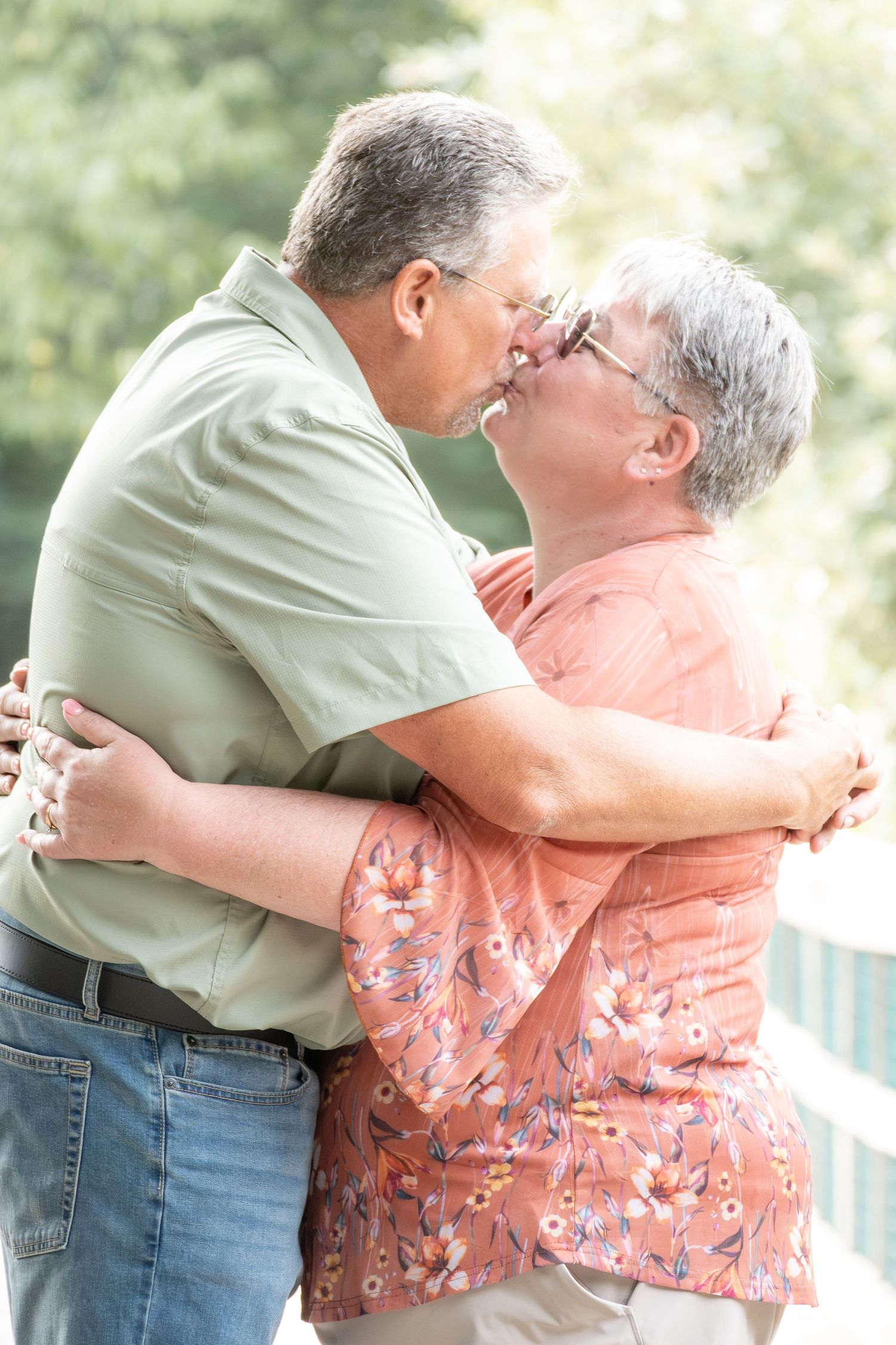 An elderly couple shares a tender kiss while embracing outdoors, surrounded by lush green trees.