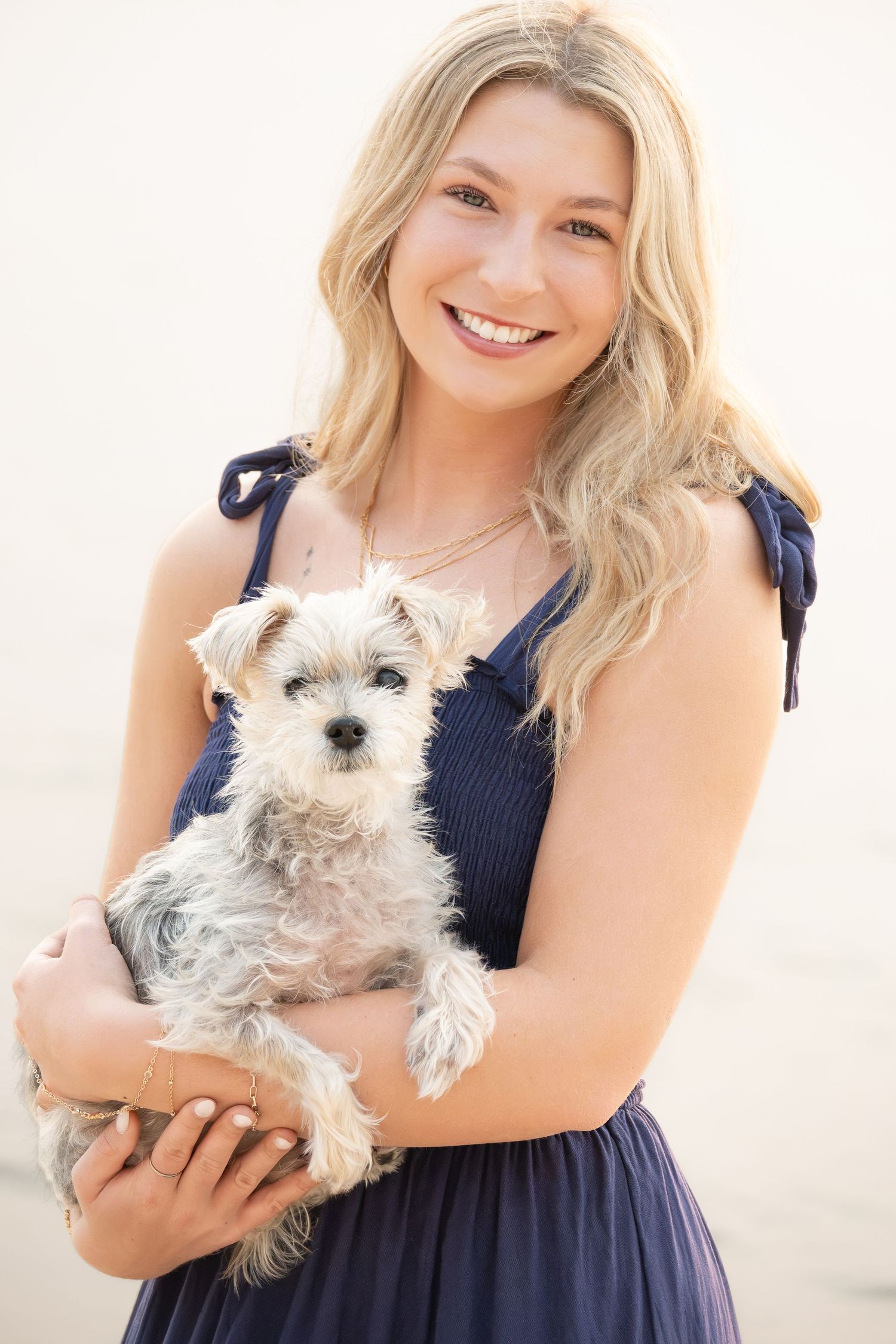 Smiling blonde woman in navy dress holding a small fluffy terrier dog outdoors.