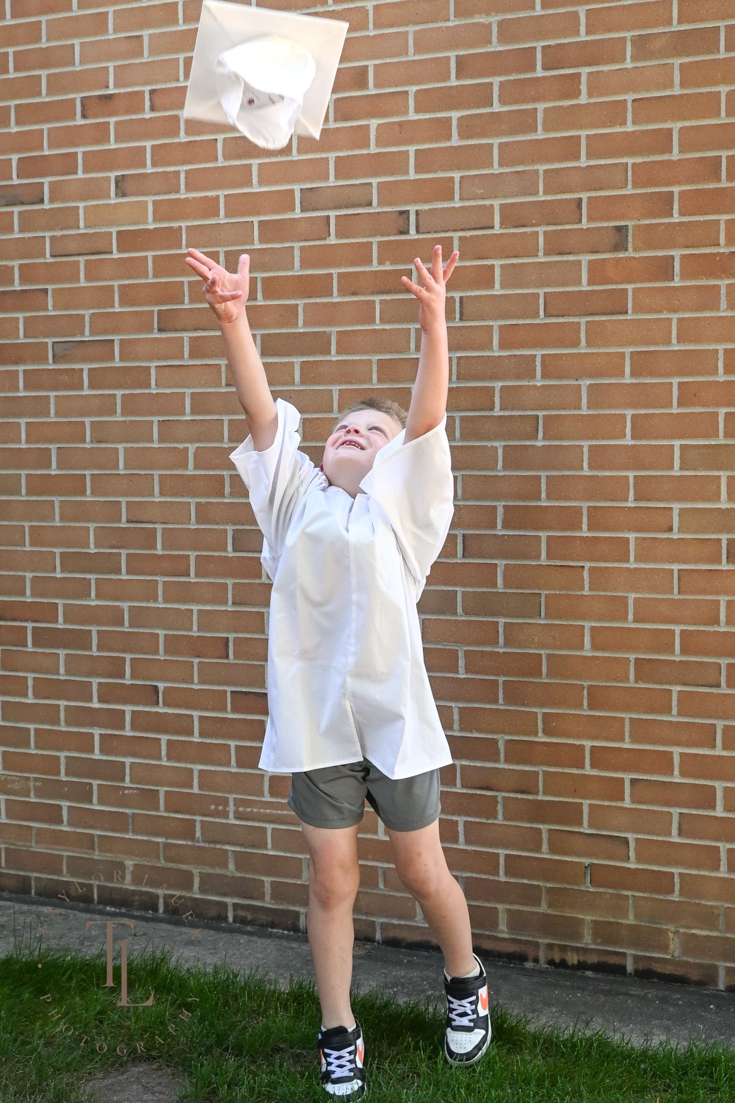 Excited young boy tossing white graduation cap in the air against a brick wall background.