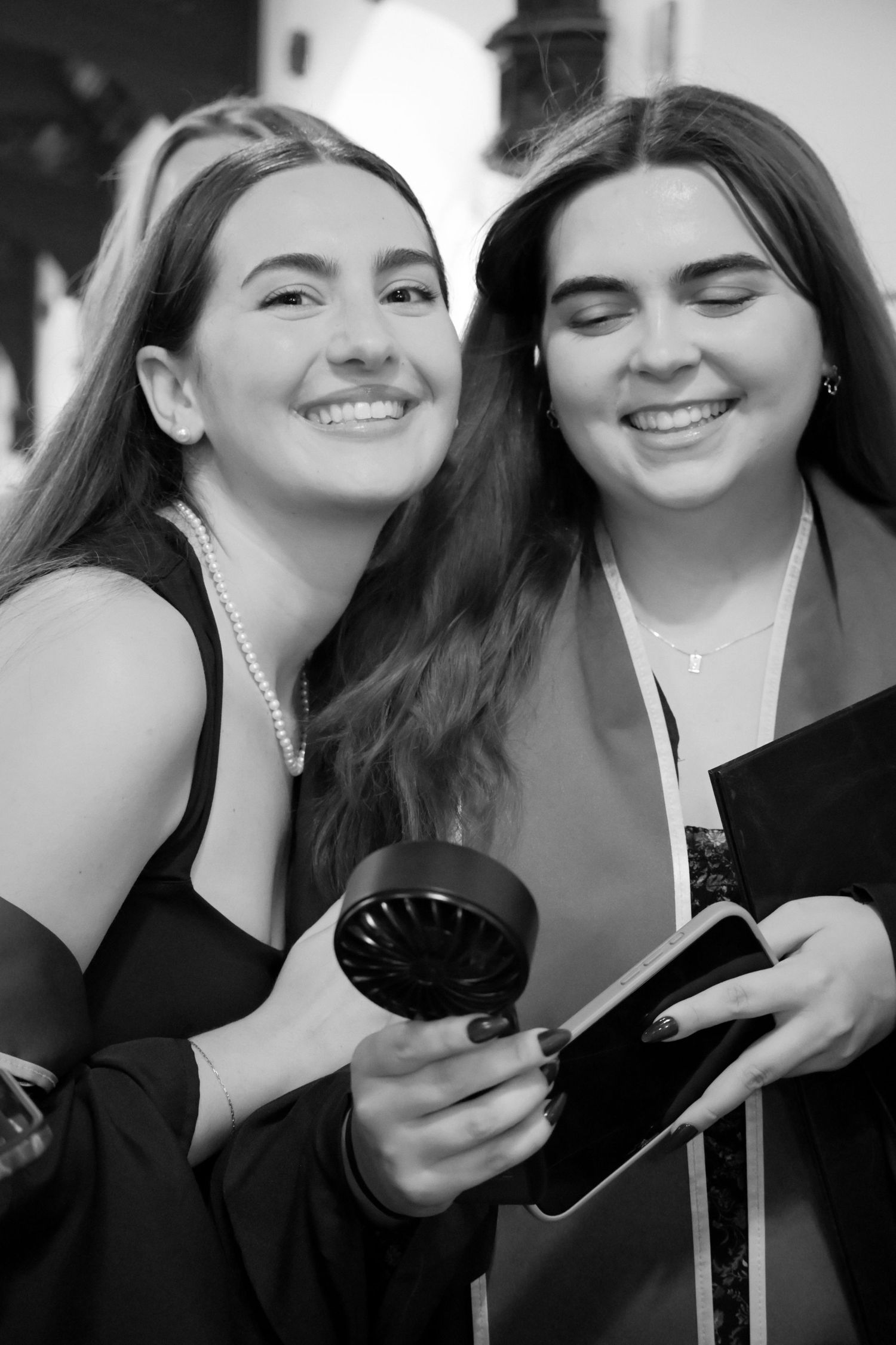 Two smiling young women in graduation attire pose together, one holding a small fan and phone.