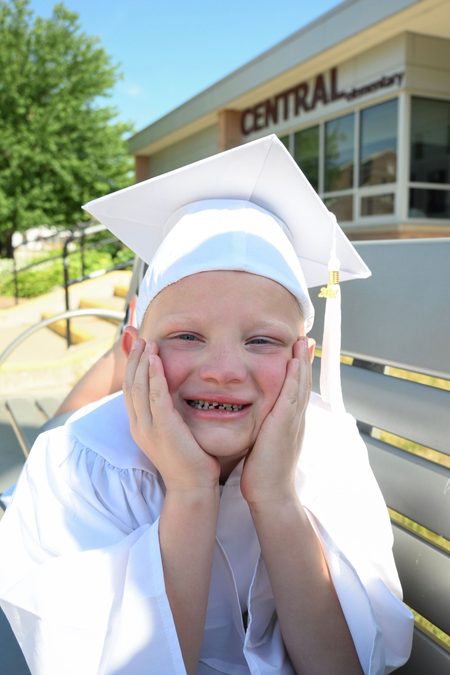 Young child in white graduation cap and gown smiling at Central Elementary School graduation ceremony.
