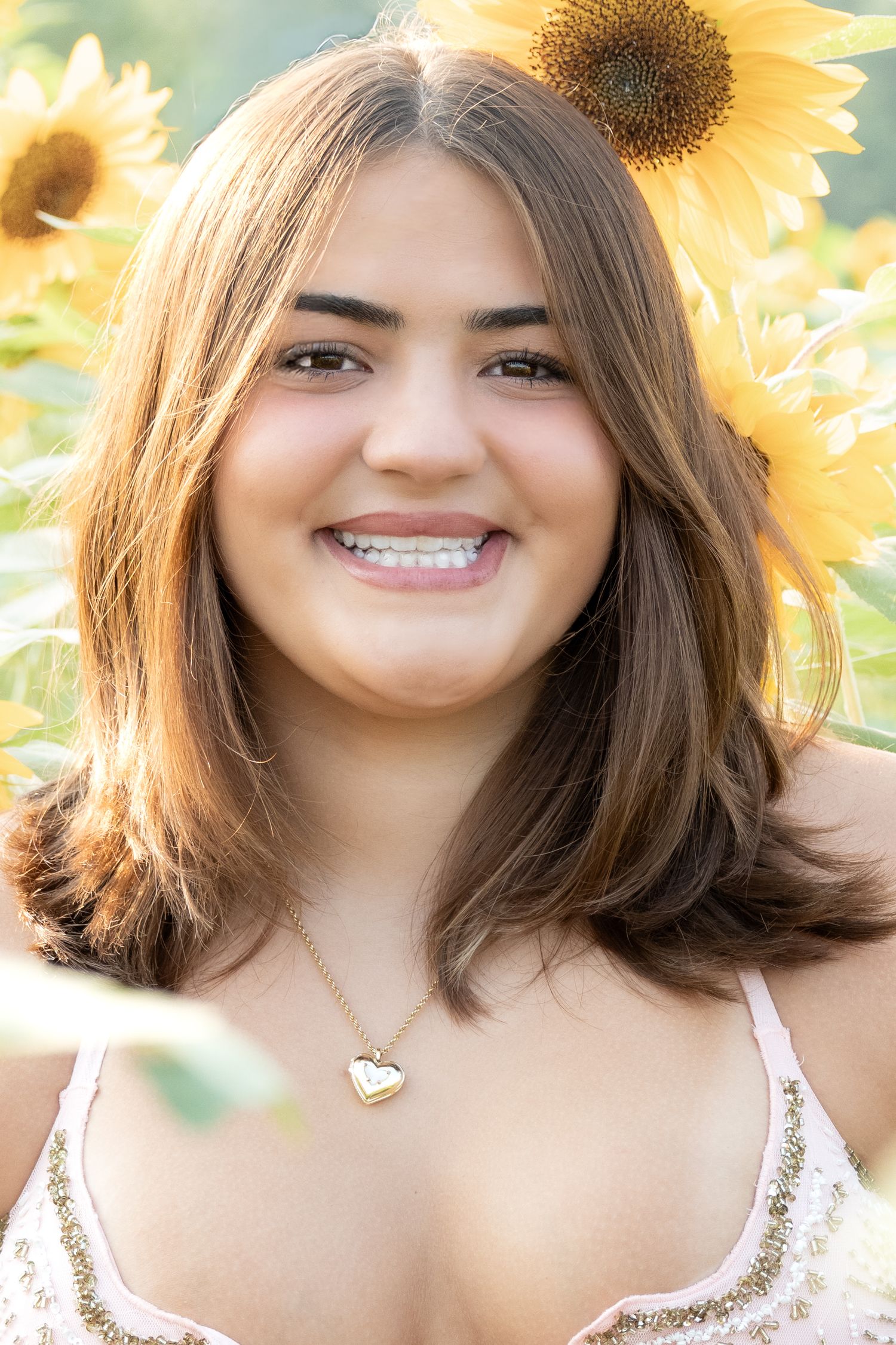 Smiling young woman with brown hair wearing a heart necklace posing in a vibrant sunflower field.
