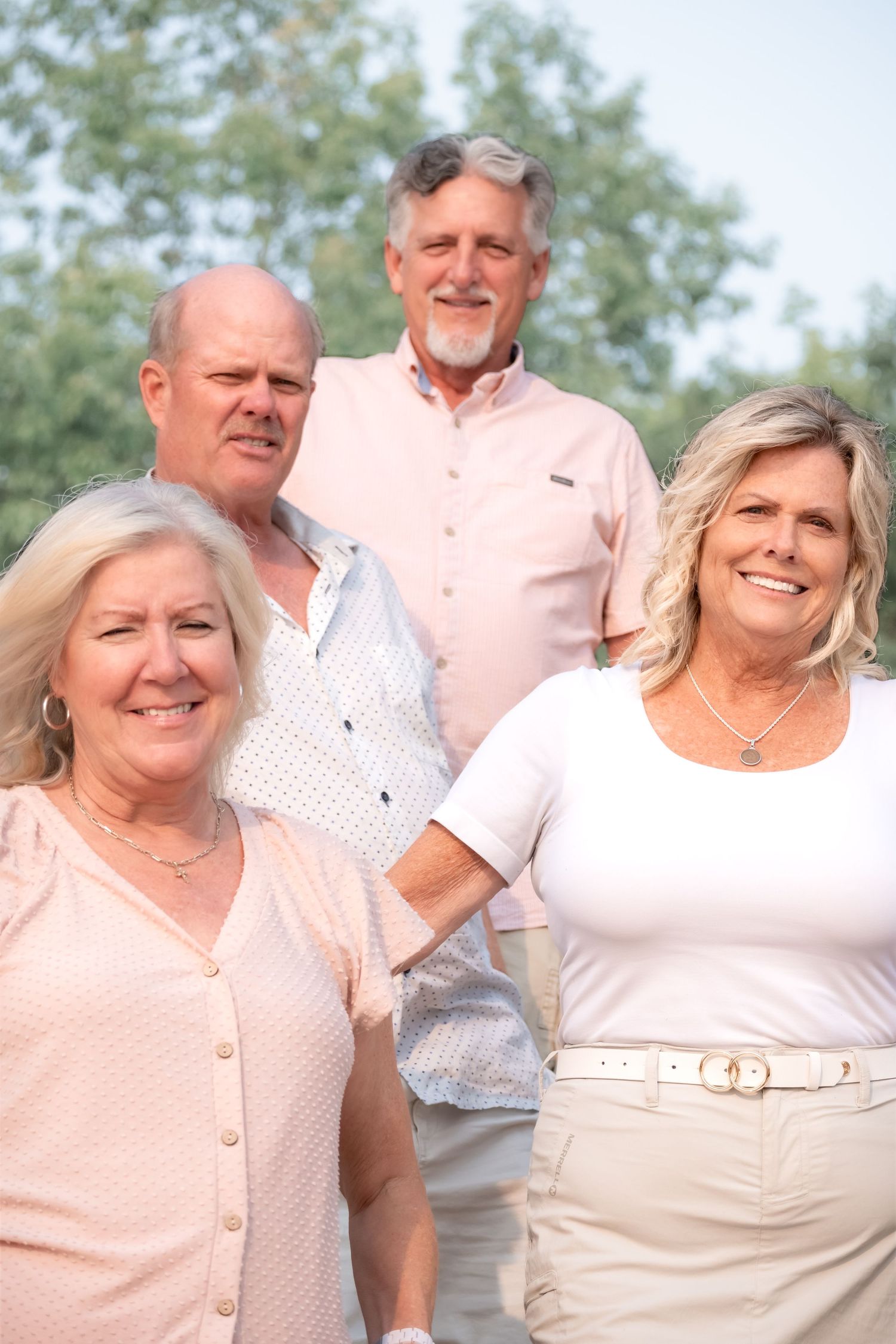 Two couples smiling outdoors in casual attire with green trees in the background on a sunny day.