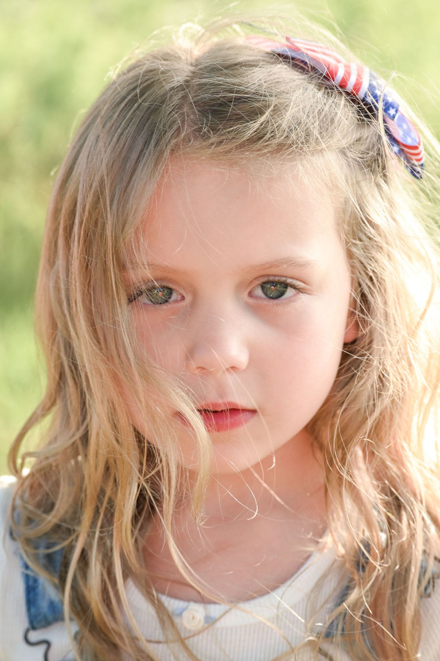 Young blonde girl with American flag hair bow poses outdoors with green background.