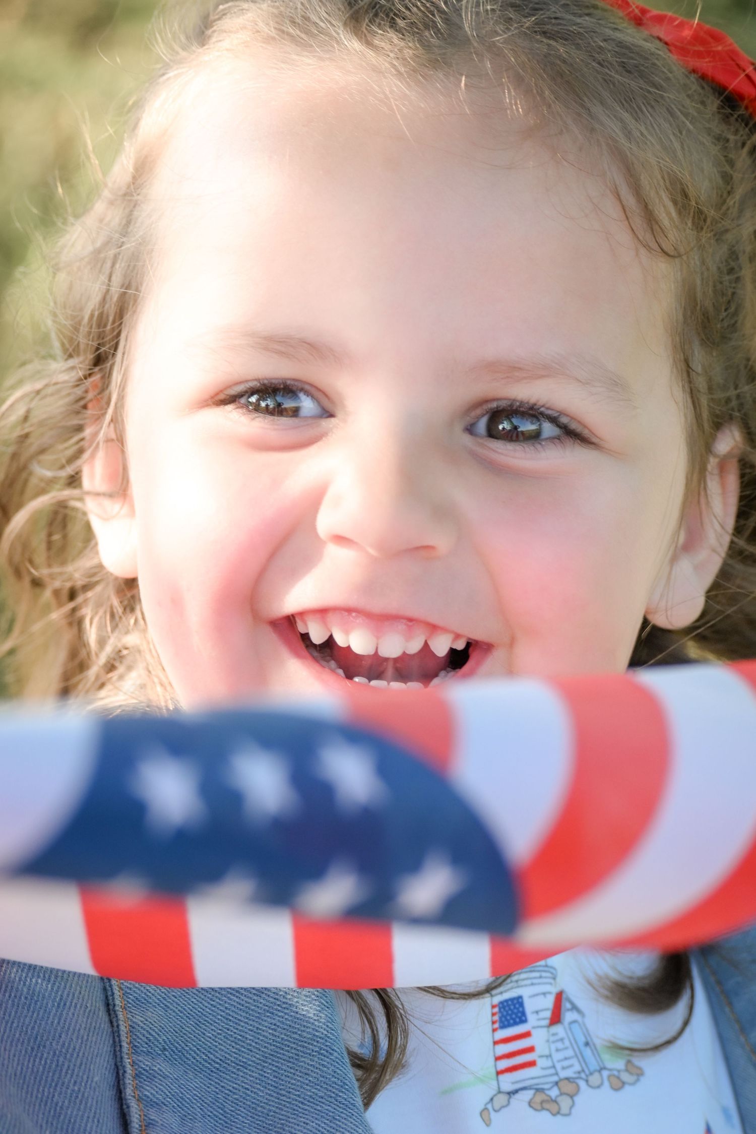 Smiling young girl holding an American flag, wearing a denim jacket outdoors on a sunny day.