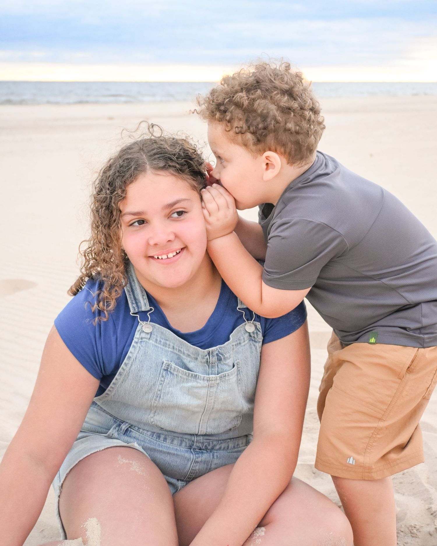 A young boy whispers into his smiling sisters ear on a sandy beach.